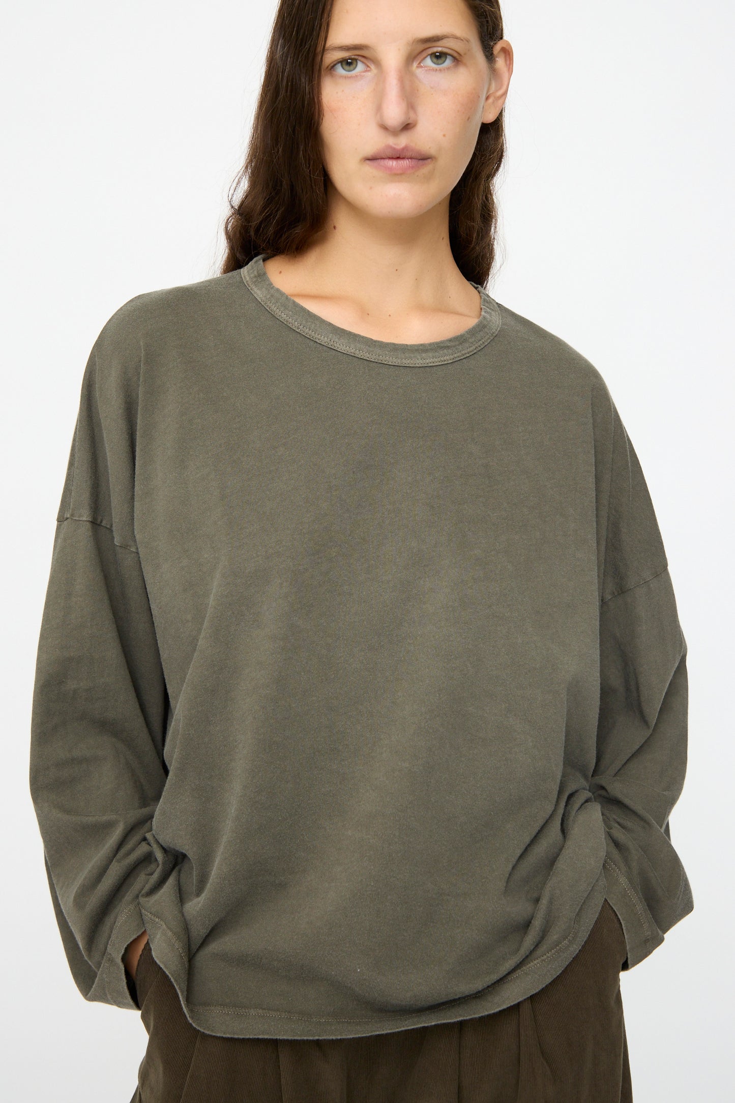 A person with long brown hair wears the Black Crane Wide Long Sleeve Tee in Slate and brown pants, standing against a plain light background.