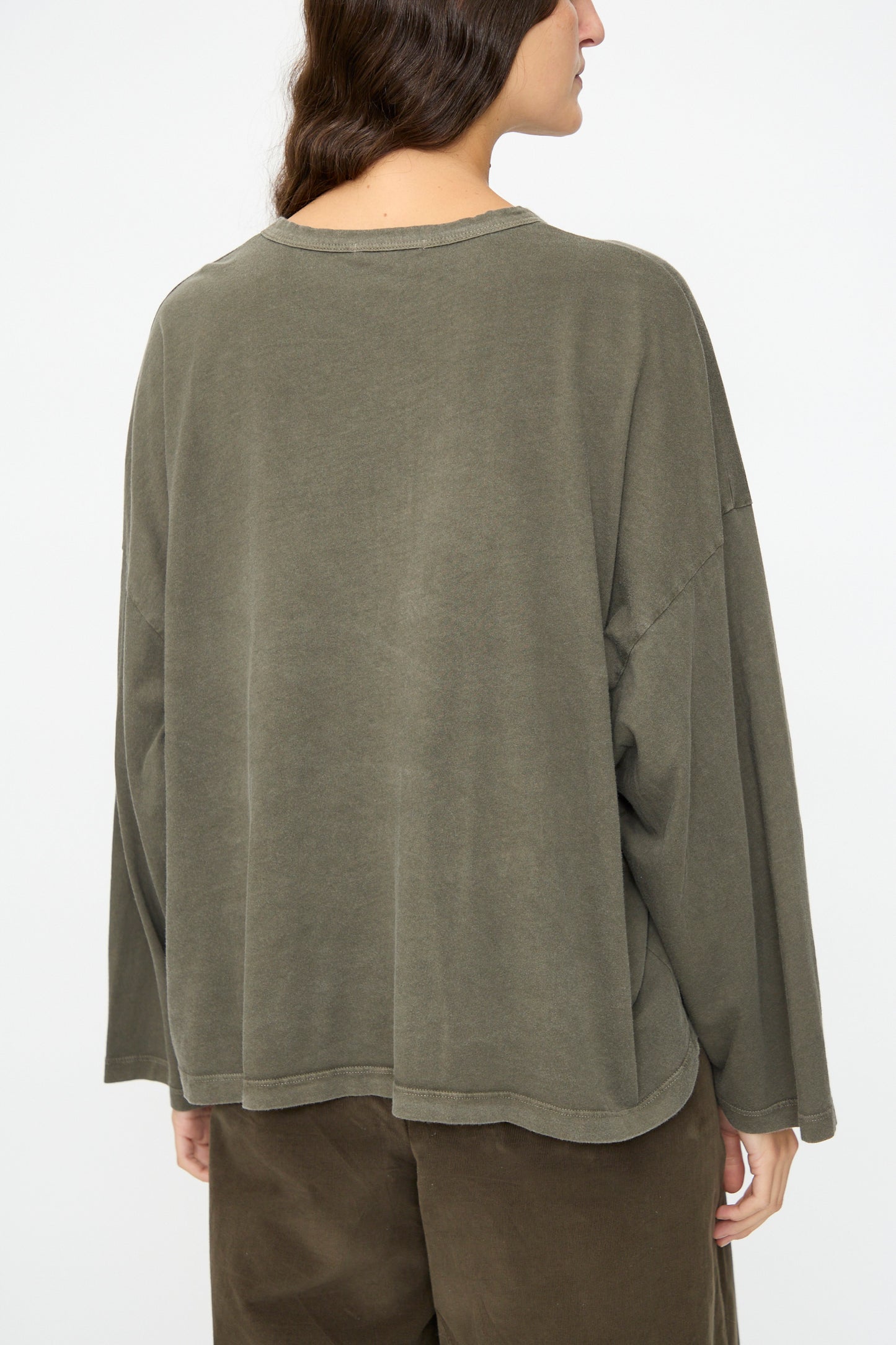 A person with long brown hair is shown from the back wearing Black Crane's Wide Long Sleeve Tee in Slate, made in the USA, paired with brown pants, against a plain background.