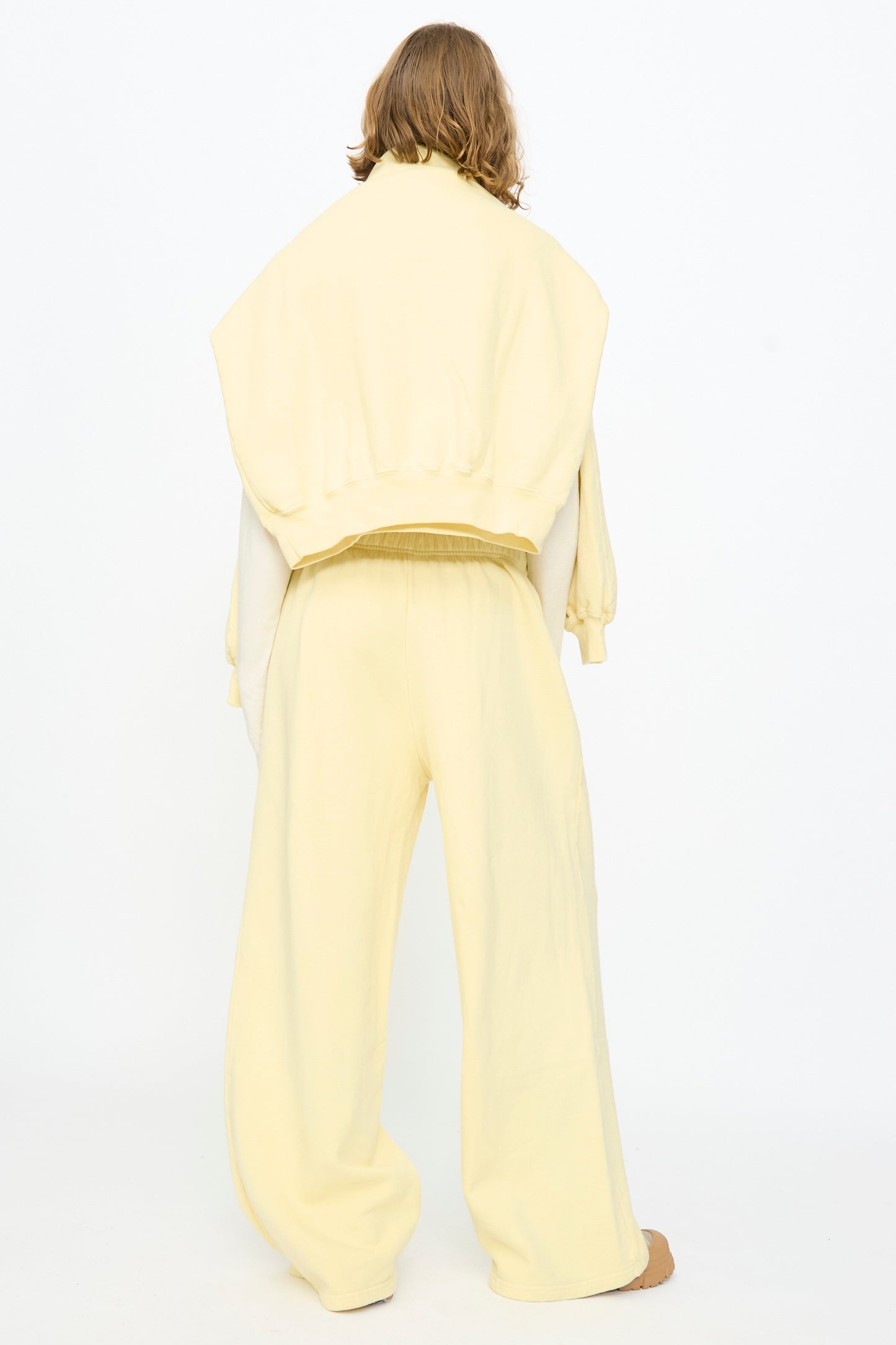 A person stands with their back to the camera, wearing Black Crane’s Wide Long Sweatpants in Custard—loose-fitting and pale yellow—with a sweatshirt draped over their shoulders, set against a plain white background.