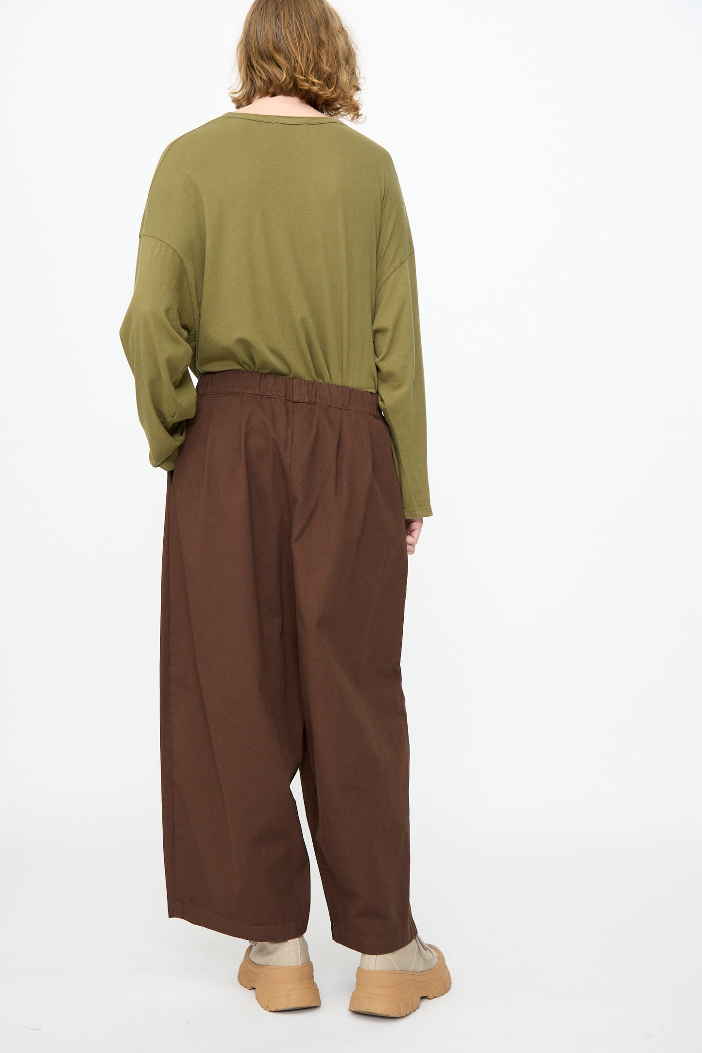 A person stands against a white background, wearing an olive green long-sleeve shirt and the Black Crane Wide Pant in Dark Cacao—loose brown cotton pants with an elastic waist—paired with beige shoes, seen from the back.
