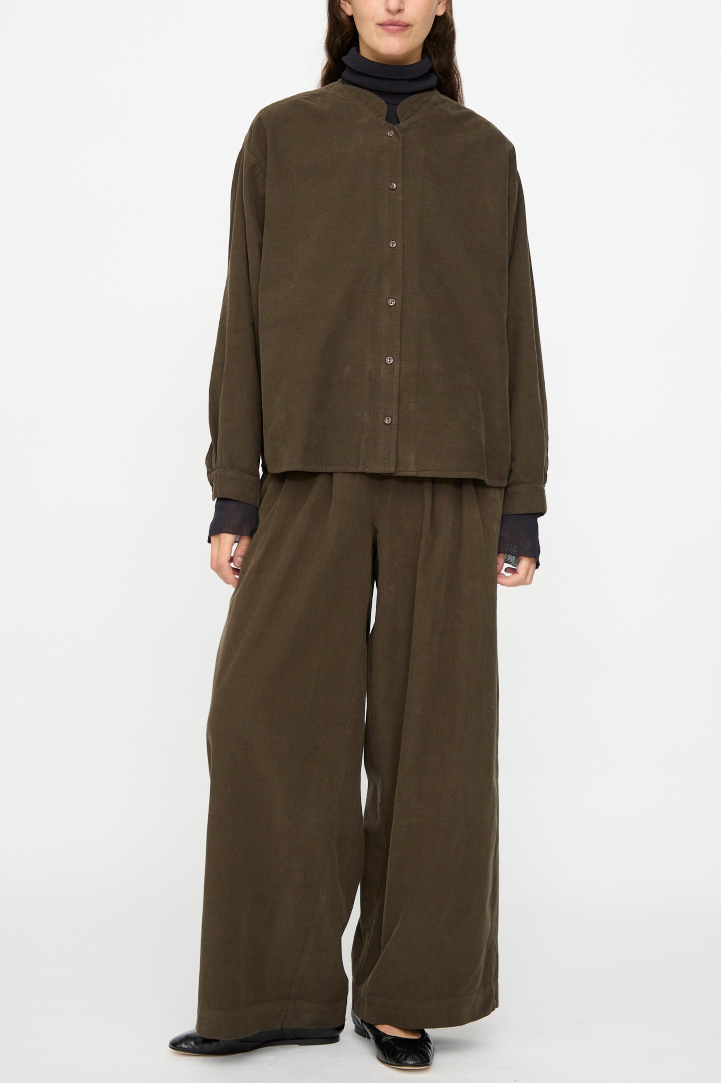 A person stands against a plain white background wearing Black Crane's Wide Pleated Pants in Dark Forest with a relaxed fit brown button-up shirt layered over a black turtleneck.