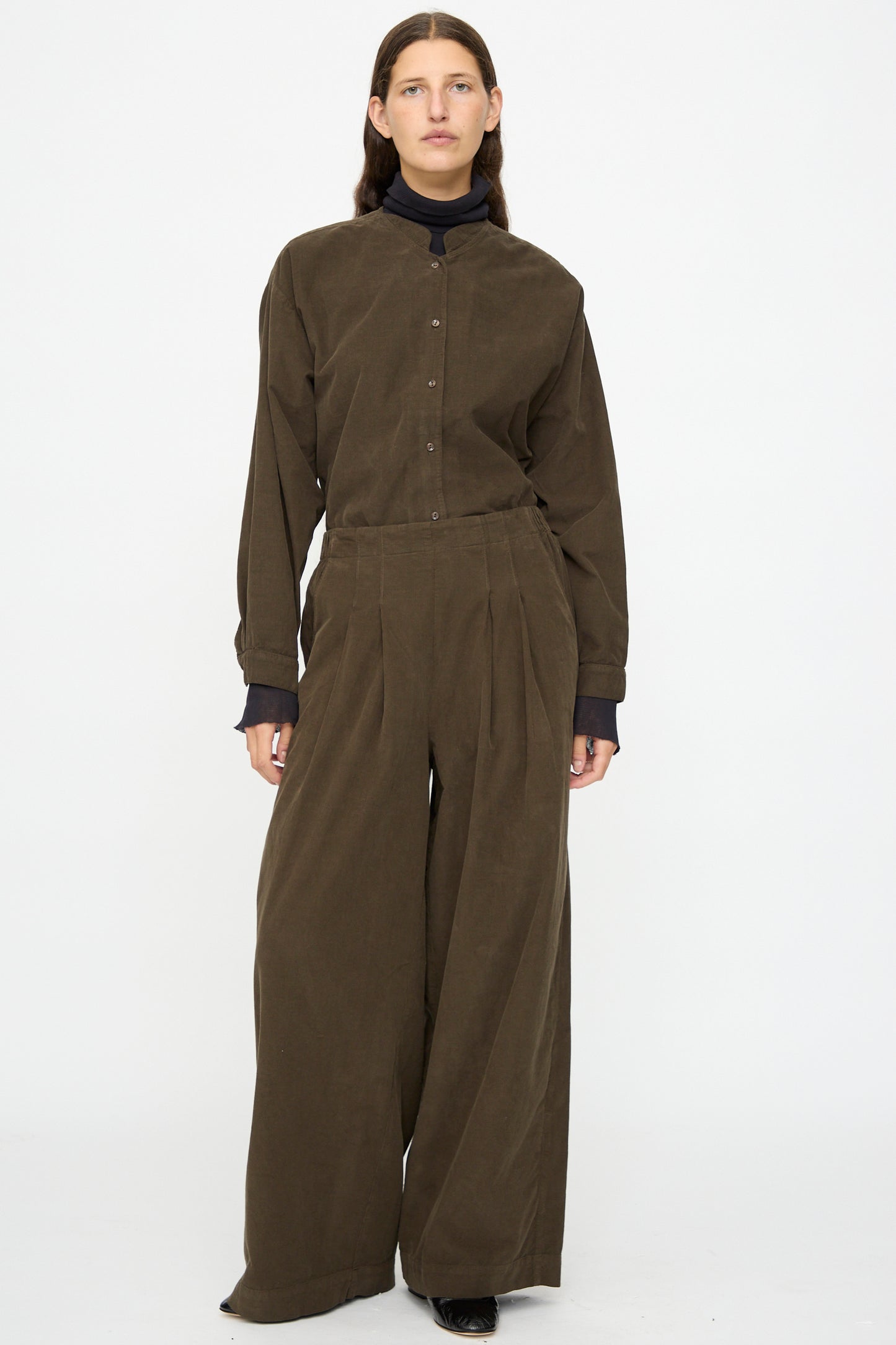 A person stands against a plain background wearing Black Crane's Wide Pleated Pants in Dark Forest with a brown relaxed-fit button-up and black turtleneck.