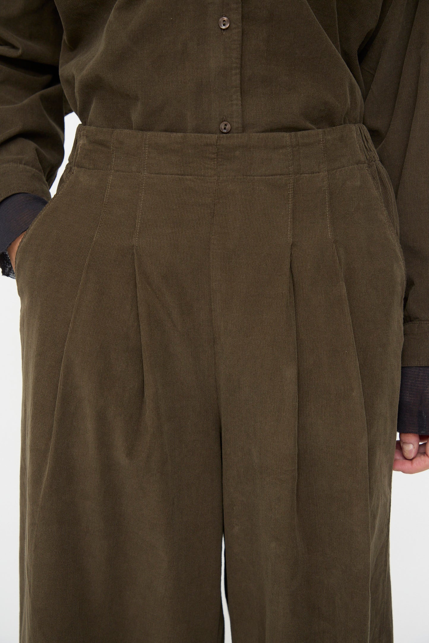 Close-up of a person wearing Black Crane Wide Pleated Pants in Dark Forest, paired with a matching button-up shirt; one hand is in their pocket.