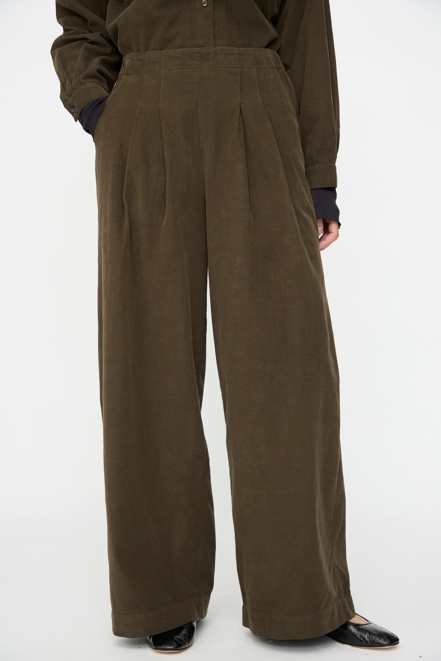 A person wears Black Crane's Wide Pleated Pants in Dark Forest—relaxed fit, high-waisted, wide-leg—with long sleeves and black pointed shoes, standing against a plain white background.