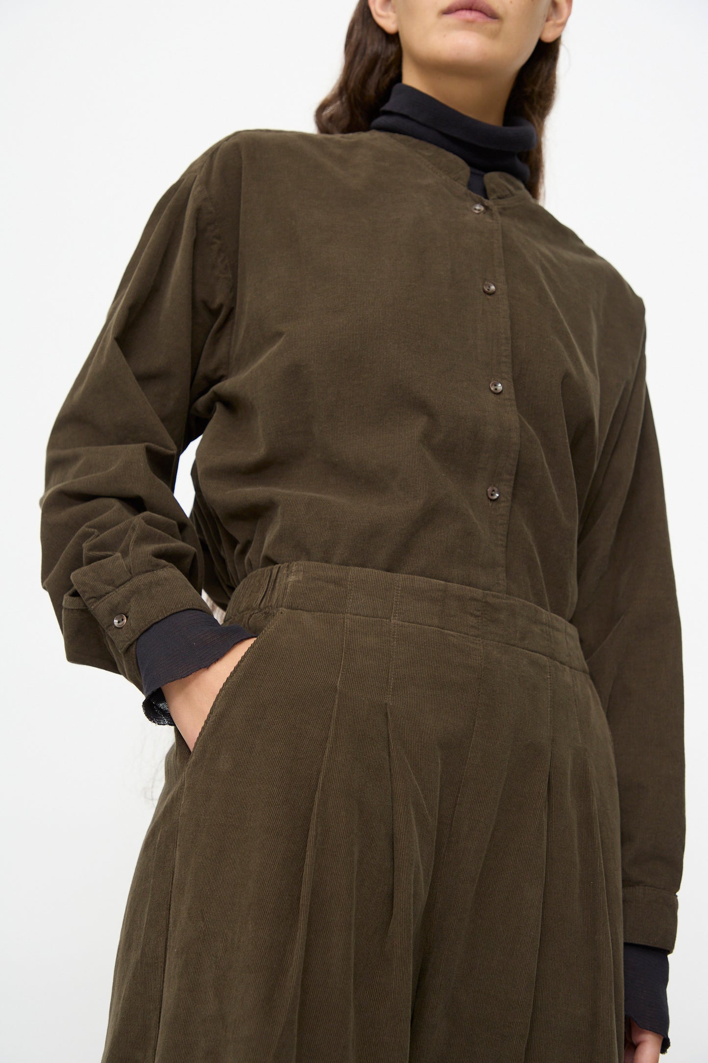 A person wears Black Crane's Wide Pleated Pants in Dark Forest paired with an olive-green corduroy shirt, one hand in their pocket, standing against a plain background.