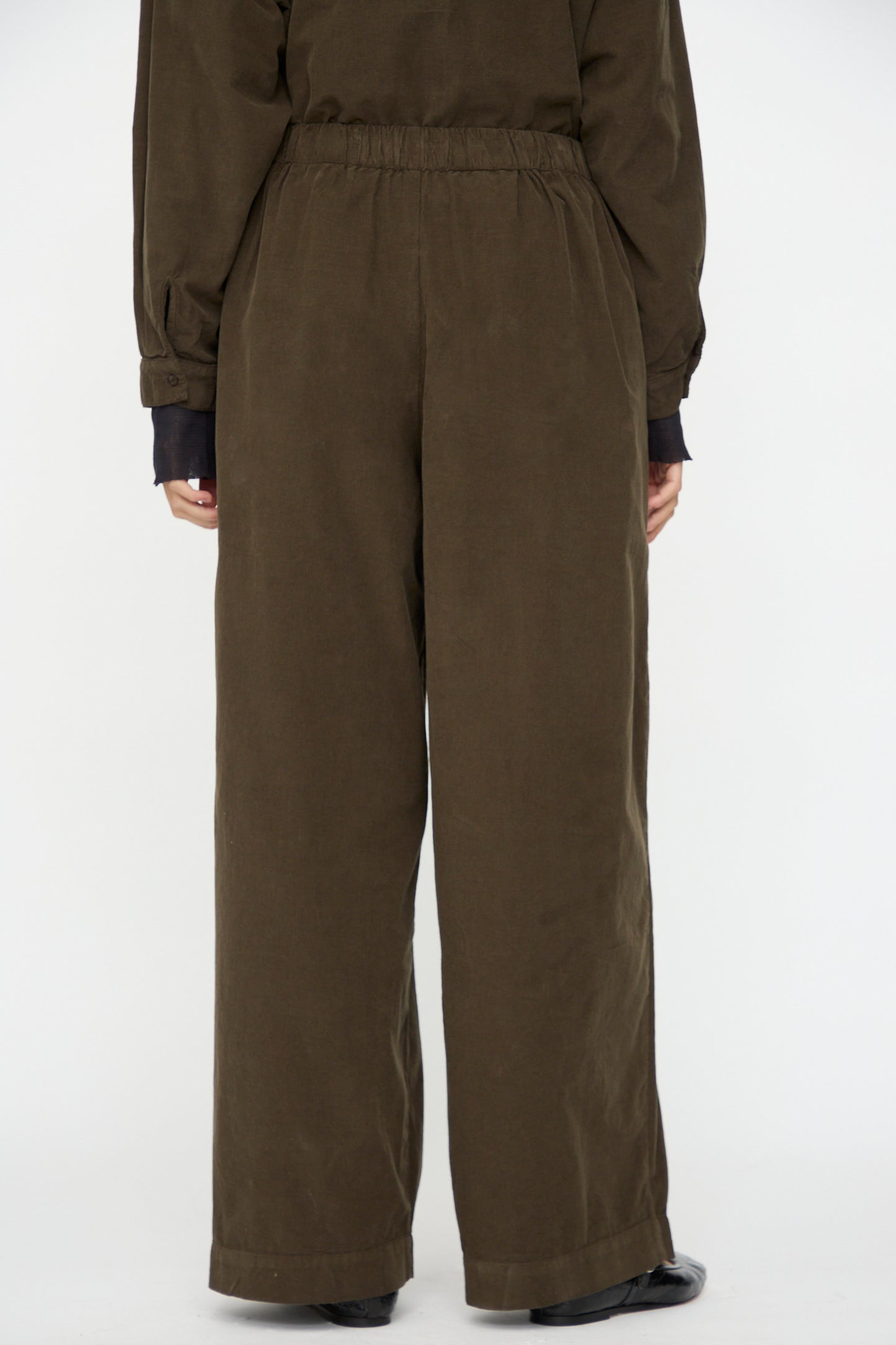 A person wears Black Crane's Wide Pleated Pants in Dark Forest with black shoes, photographed from the back against a plain white background.
