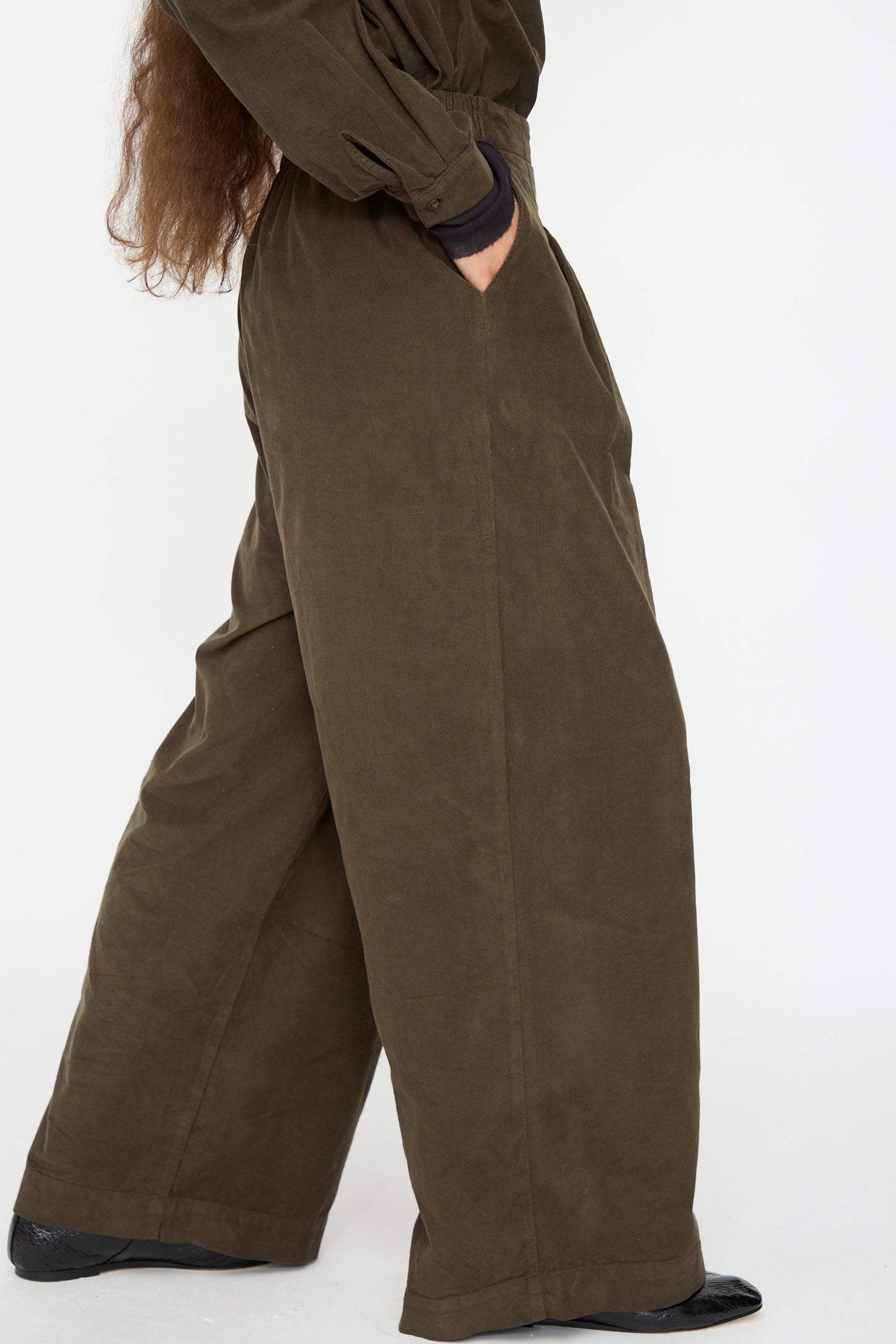 Model wears Black Crane's Wide Pleated Pants in Dark Forest, a relaxed fit, wide-leg style with hands in pockets, standing against a plain white background.