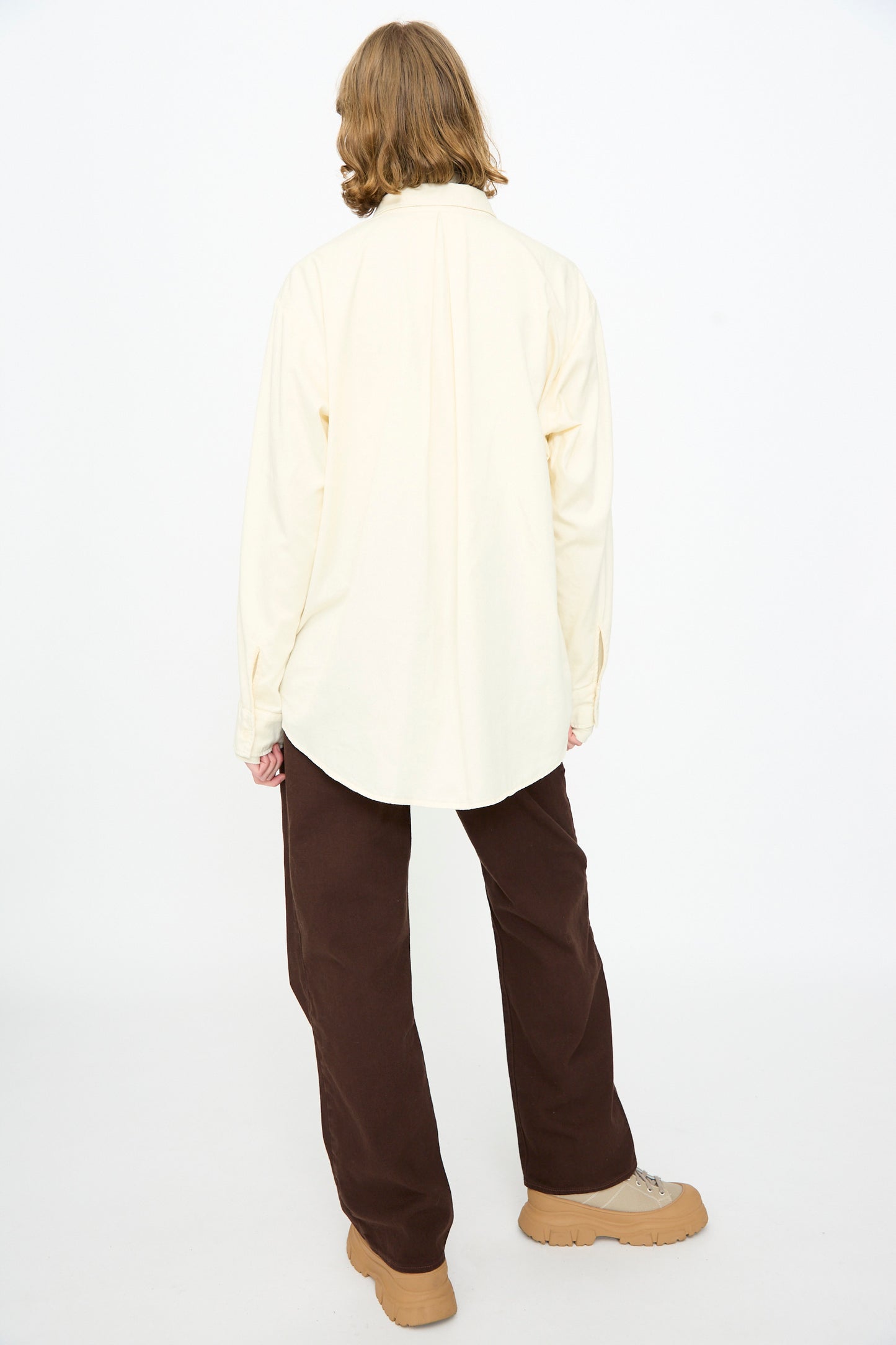 A person with shoulder-length hair stands facing away, wearing the Black Crane Work Shirt in Natural with brown pants and tan shoes against a white background.