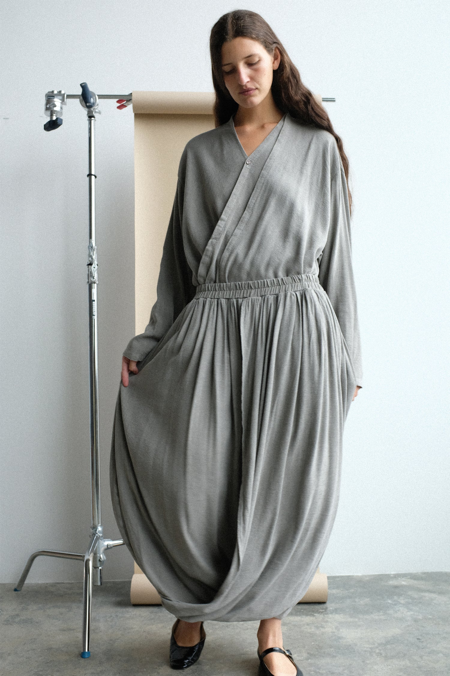 Woman wearing a long, light gray top and skirt standing in front of a tan seamless.