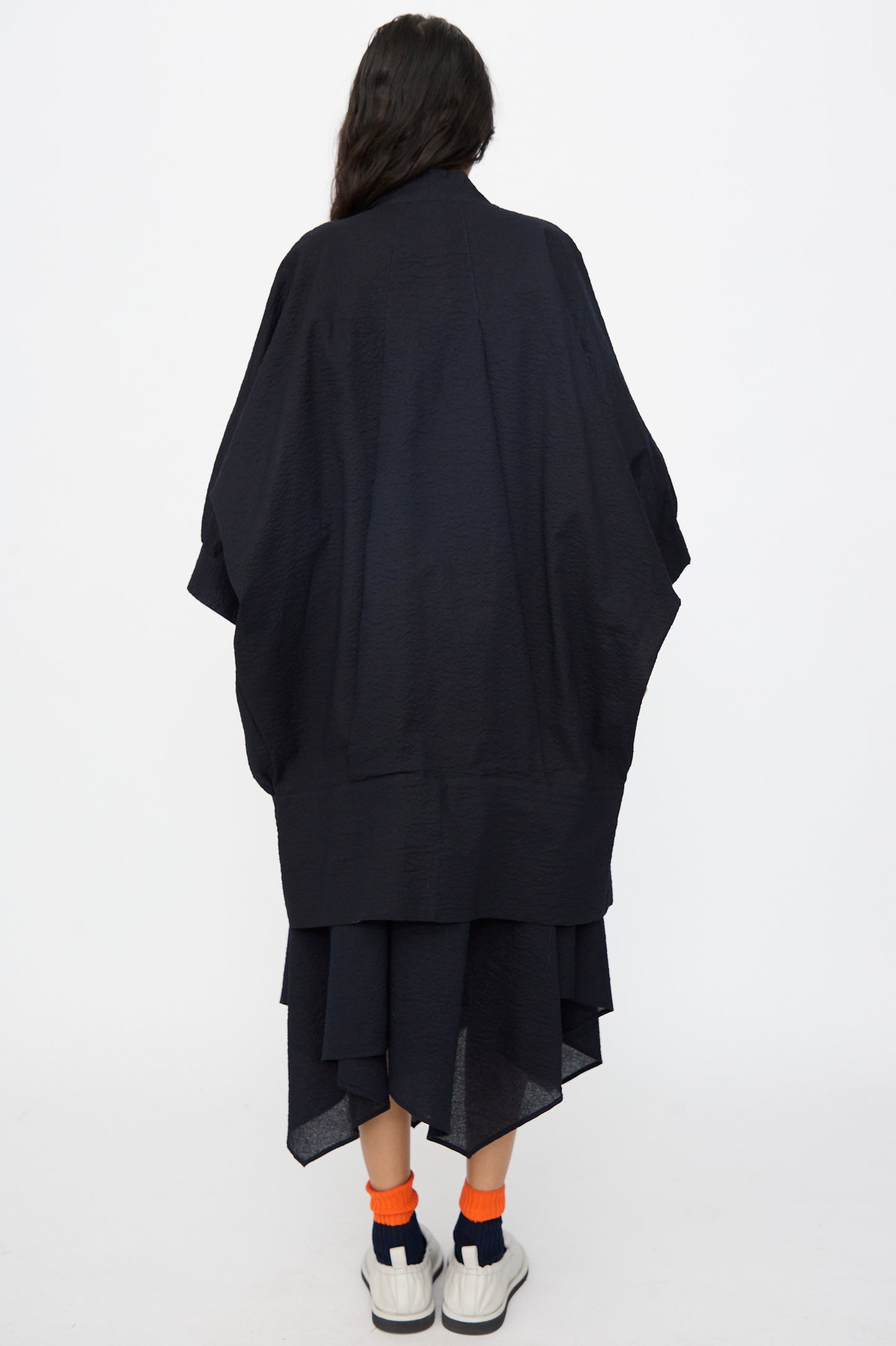 A person with long dark hair faces away, wearing the Black Crane Textured Cotton Spoon Jacket in Midnight, paired with white sneakers and orange and navy socks, set against a plain white background.