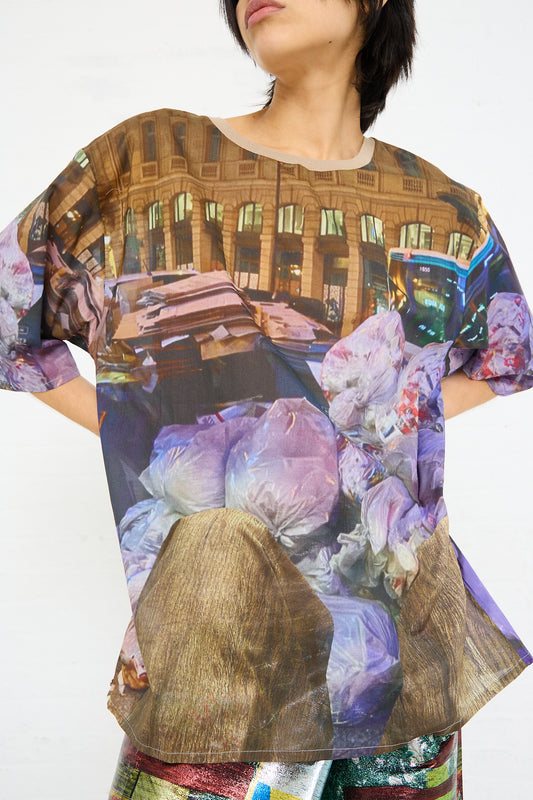 A person wearing a lightweight Bless Cotton No. 77 Holiday Paris Strike Tee in Print with a graphic print of Paris urban scenery and trash bags.
