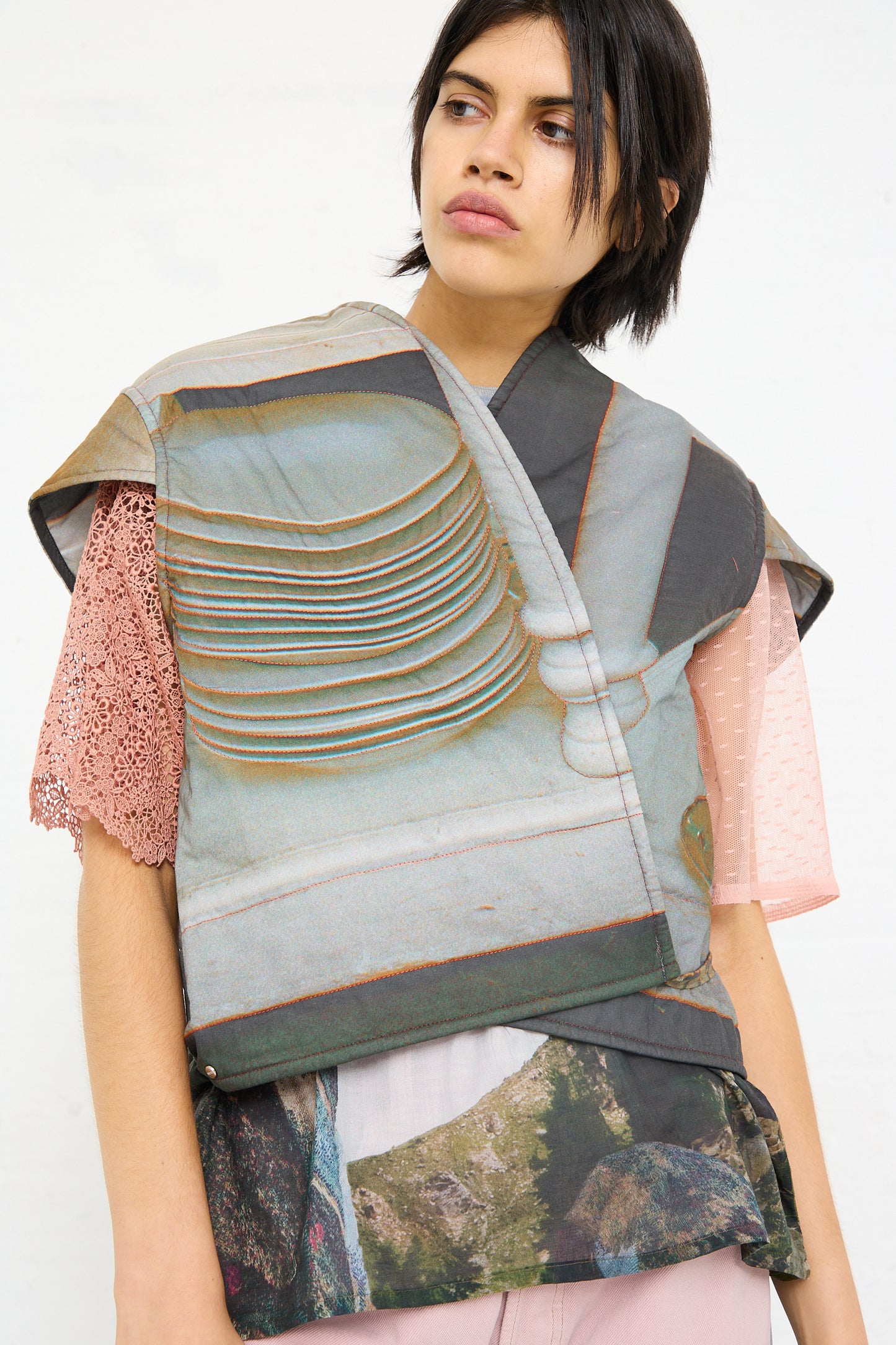 A person wearing an avant-garde, multilayered and textured outfit, highlighted by an oversized Cotton No. 77 Summer Living vest in print from the Bless luxury brand, with a contemplative expression.