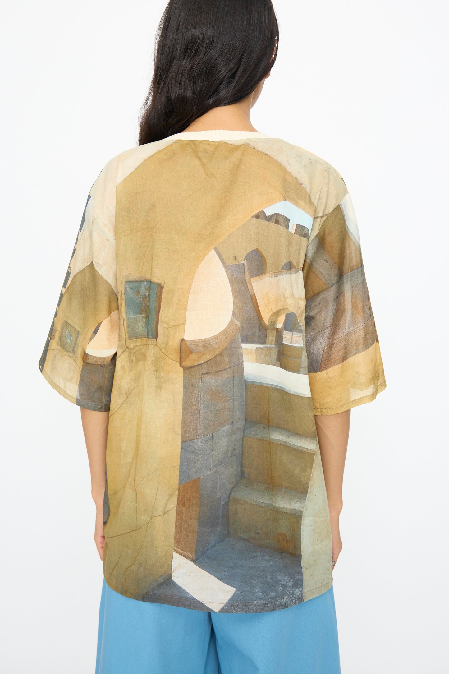A person with long dark hair, seen from the back, wears the Bless Holidayjaipurobservatory Tee in Print—an oversized, boxy fit T-shirt with an architectural graphic—paired with blue pants.