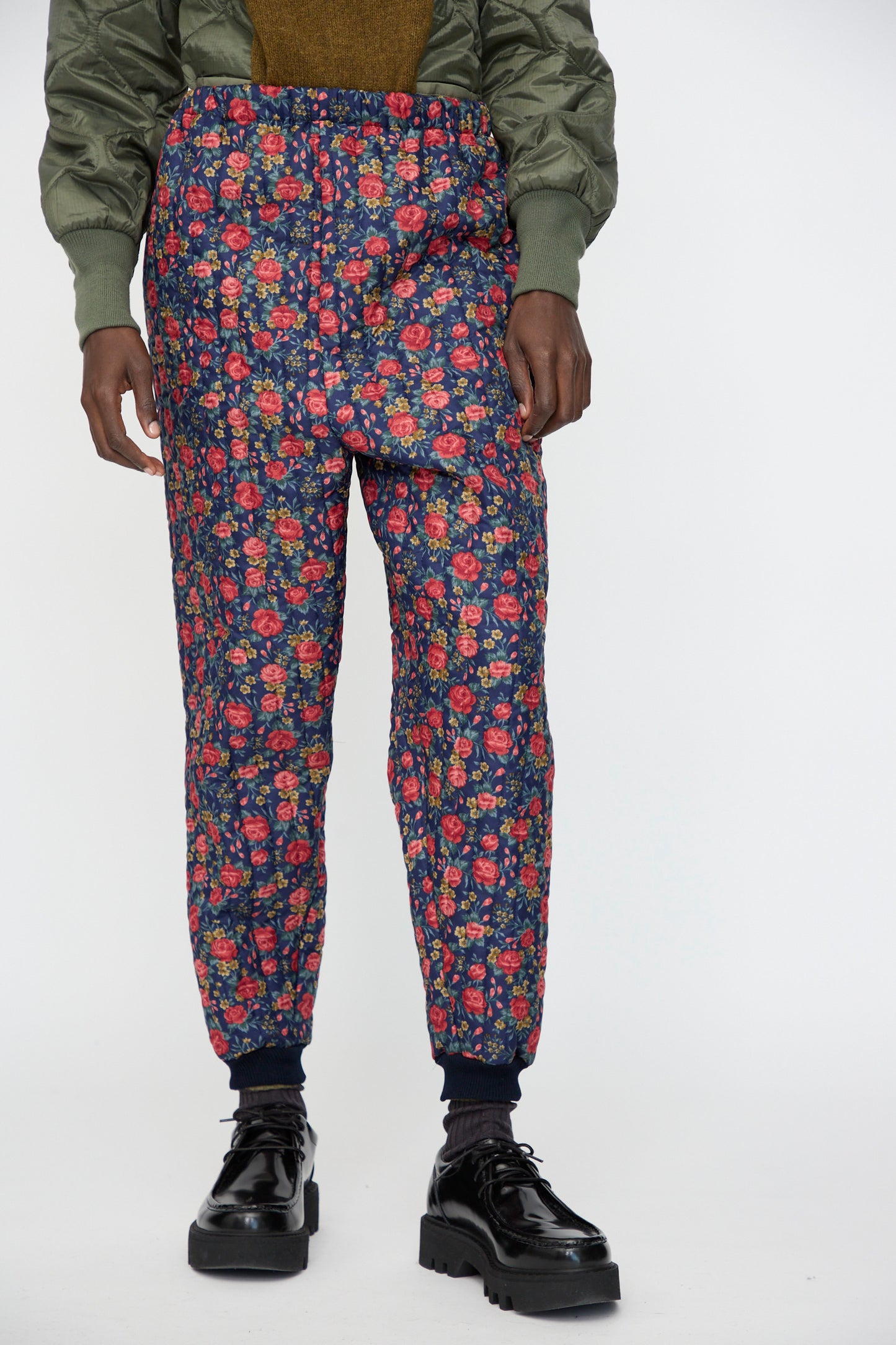 A person wearing Bless No. 08 Monpe Pant in Floral Print I, a green quilted jacket, and black platform shoes, standing against a white background.