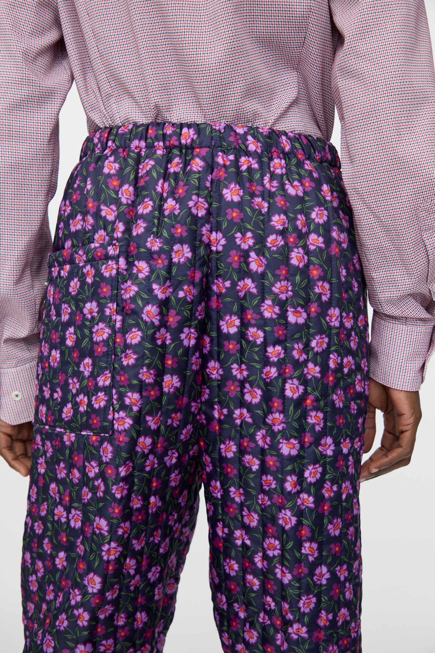 A person seen from the back wears a light red checkered shirt and Bless’s No. 08 Monpe Pant in Floral Print II, featuring a high-waisted fit with a pink and purple floral pattern.