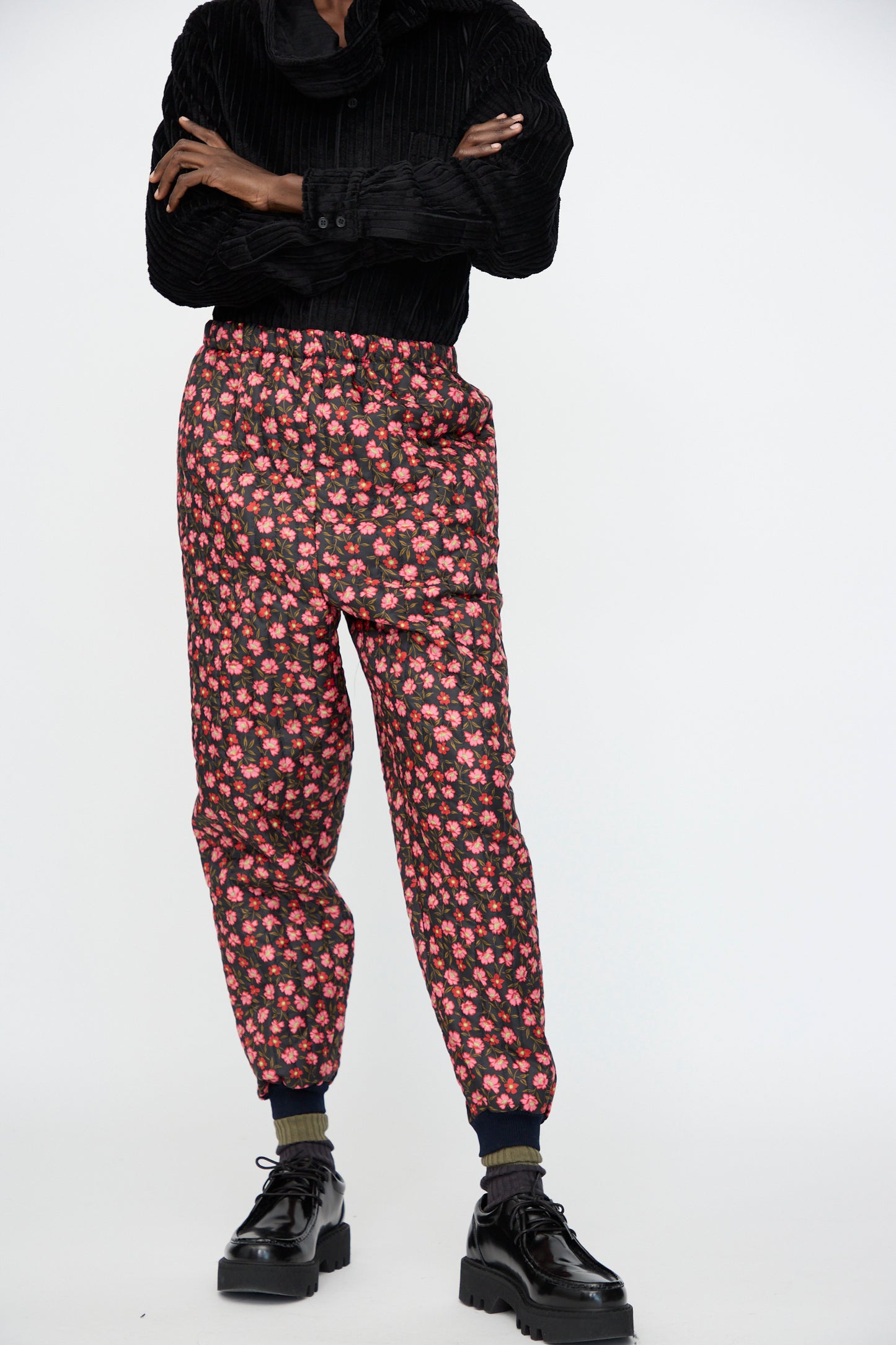 A person stands with arms crossed against a plain background, wearing a black textured shirt, Bless No. 08 Monpe Pant in Floral Print IV, dark socks, and black platform shoes.