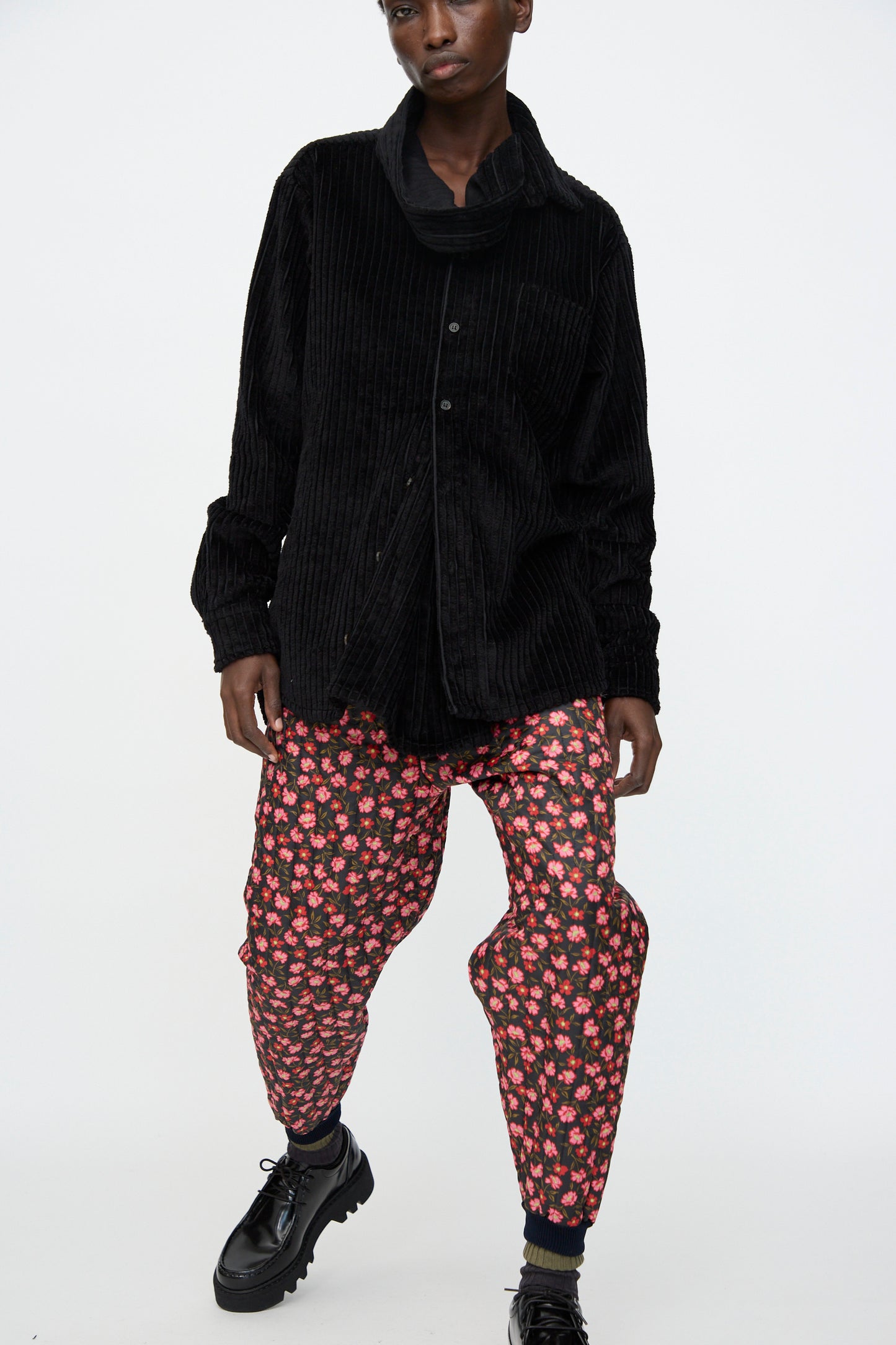 A person wears a loose black corduroy shirt, Bless No. 08 Monpe Pant in Floral Print IV, and black shoes while standing against a plain white background.
