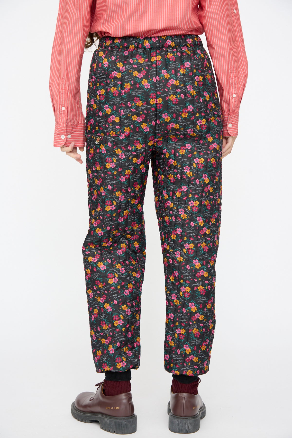 Back view of a person wearing black quilted floral No. 08 Monpe Pants in Floral Print IV and a red and white striped button-down shirt.