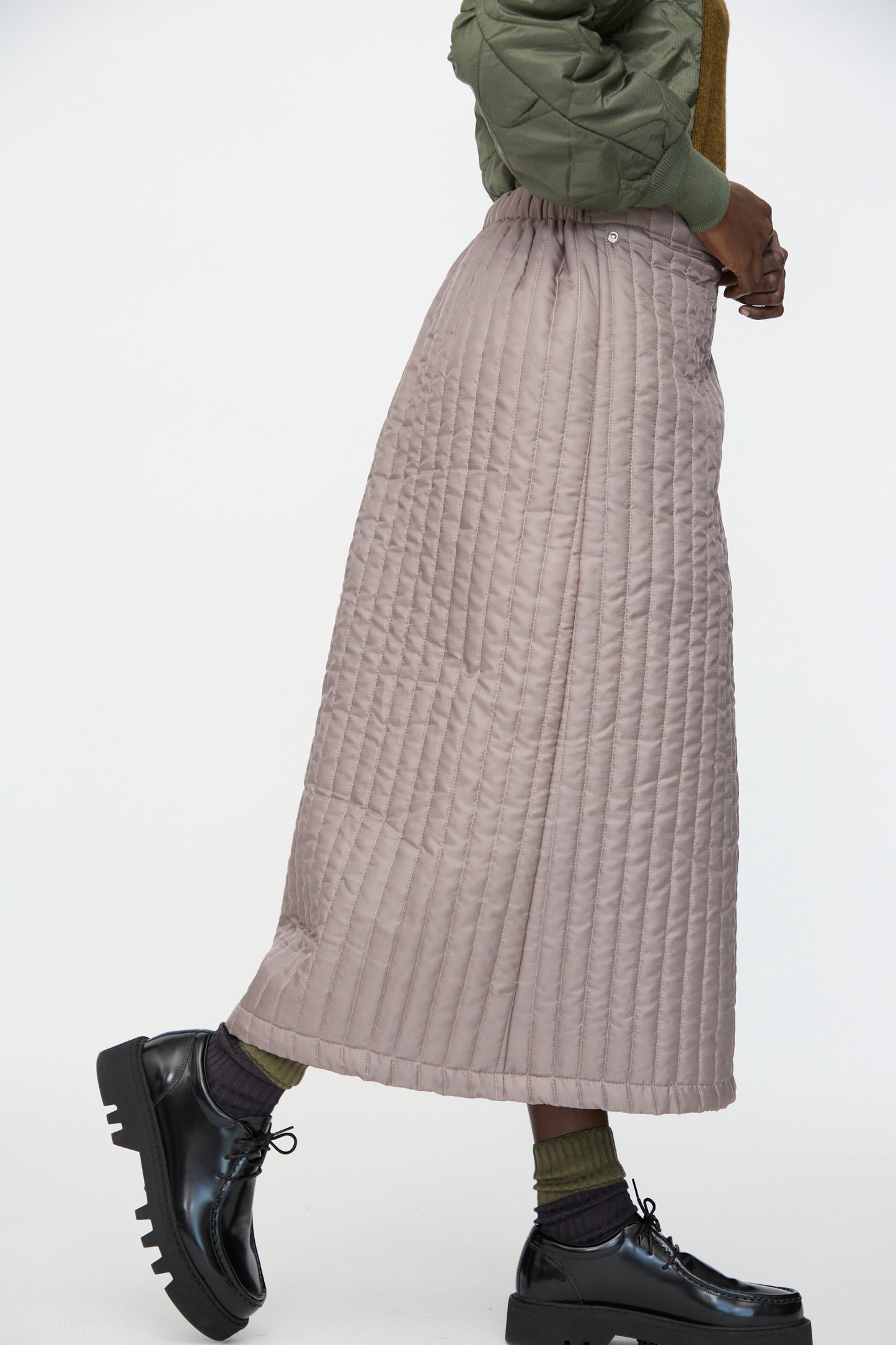 A person wearing the Bless No. 08 Monpe Skirt in Beige, an olive green jacket, black platform shoes, and green socks walks against a plain white background.