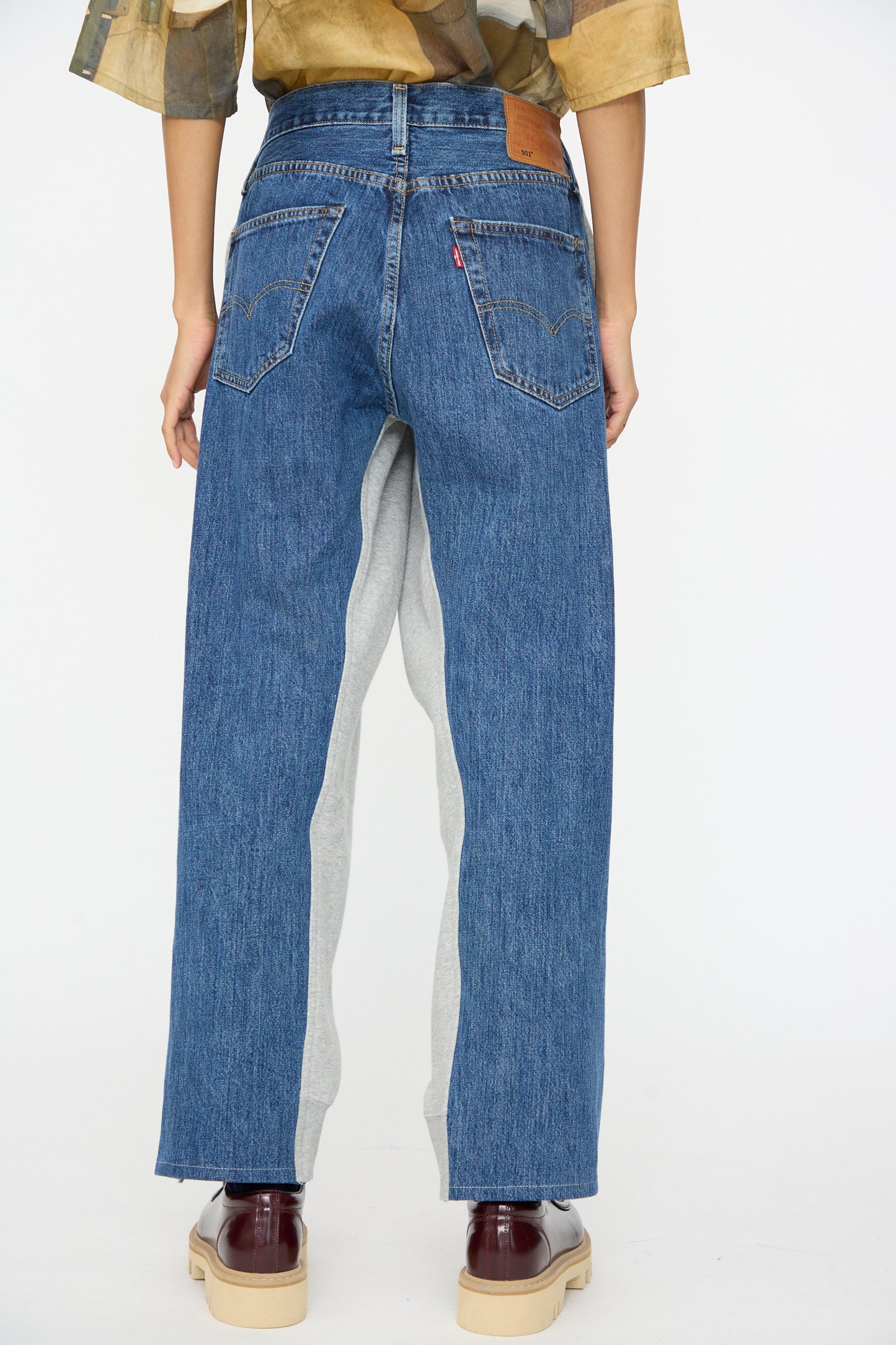 A person is seen from the back wearing Bless No. 61 Overjoggingjeans in light grey and denim blue, featuring a relaxed, straight-leg fit and paired with burgundy shoes.
