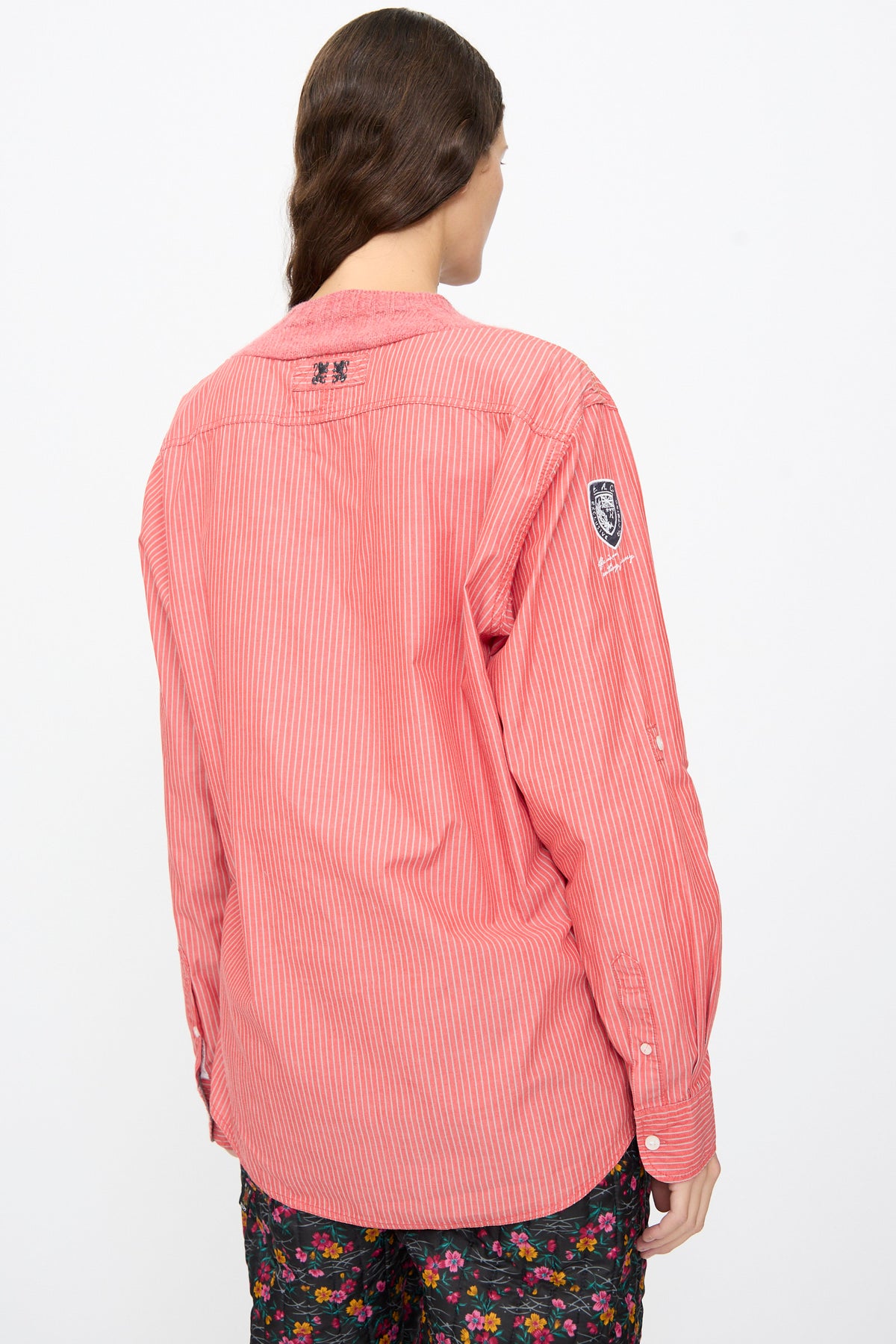 Back view of a coral pink striped Bless No. 68 Frontinsertpullover in Unique II with thin white vertical stripes, a small crest patch on the left sleeve, and black embroidery on the upper back.