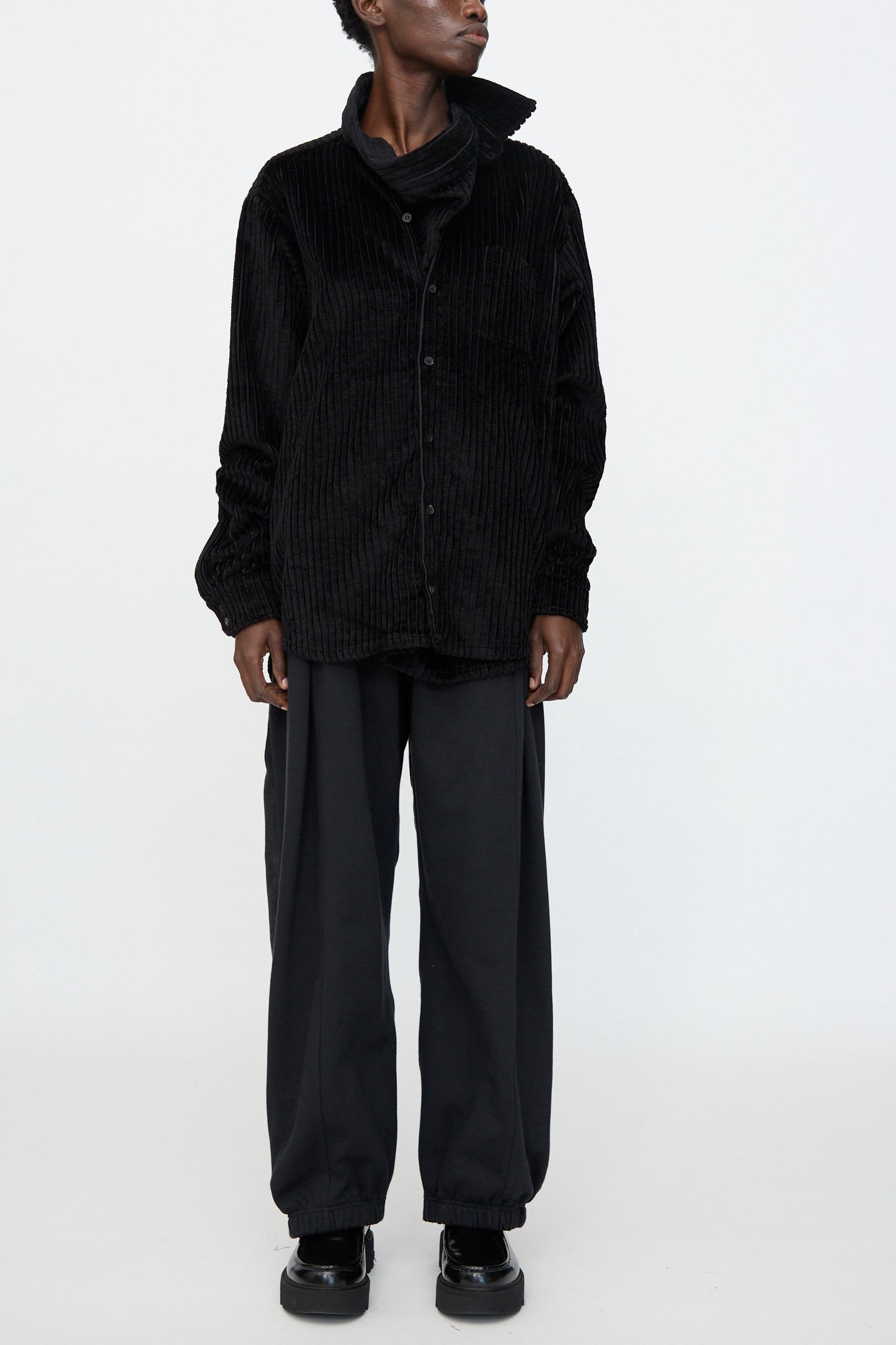 Wearing the Bless No. 68 Neckerchiefer in Black Corduroy, paired with loose dark pants and black shoes, this person stands confidently against a plain white background.