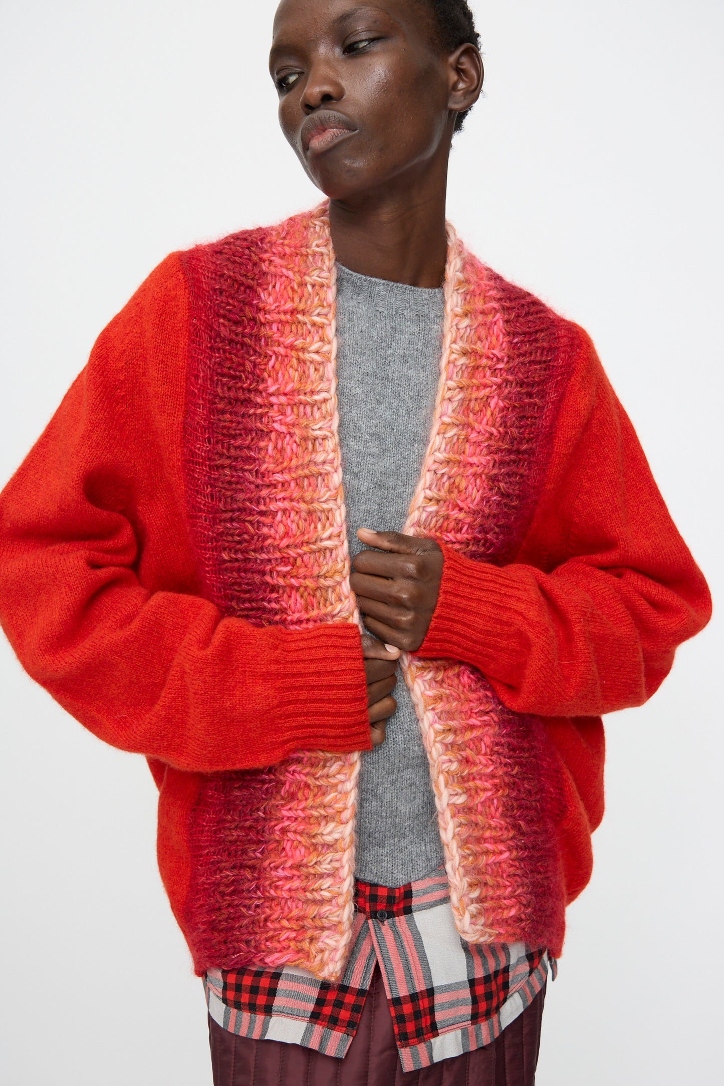 Model wearing the Bless No. 70 Passencardigan in Scarlet, a red and pink hand-knitted open cardigan sweater, layered over a gray top and plaid shirt, standing against a plain white background.