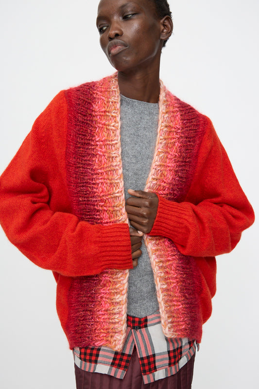 Model wearing the Bless No. 70 Passencardigan in Scarlet, a red and pink hand-knitted open cardigan sweater, layered over a gray top and plaid shirt, standing against a plain white background.
