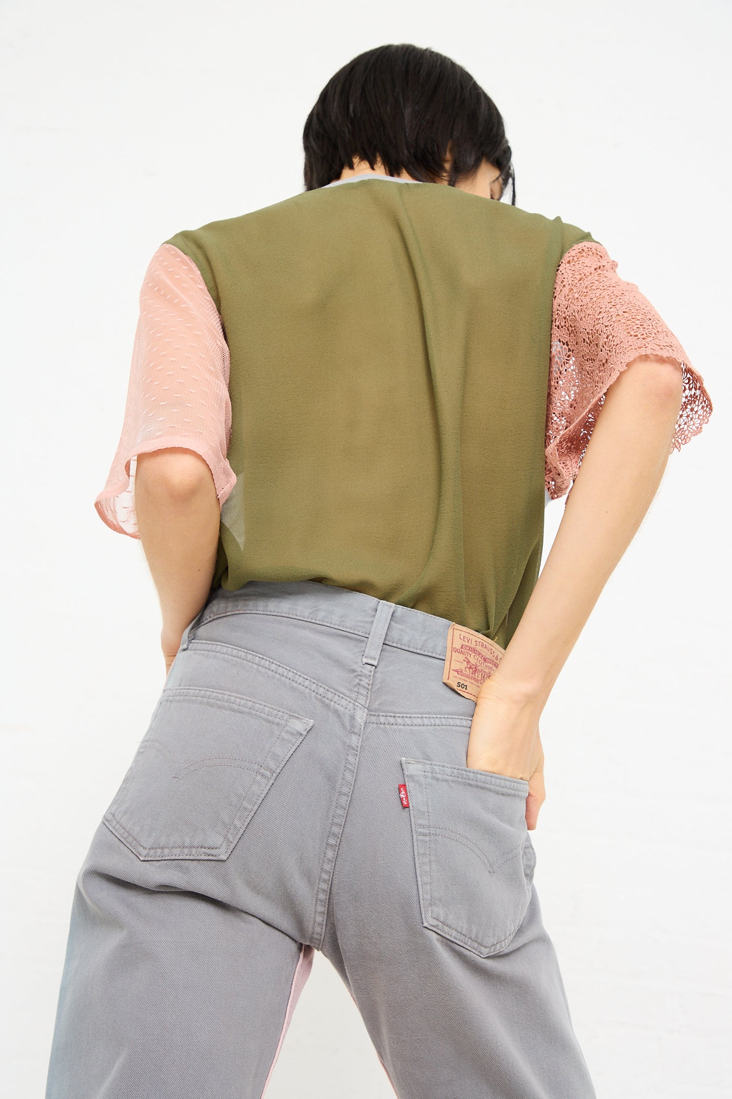 A person seen from behind wearing a green and pink top and No. 73 Jeanspleatfront in Pink/Grey mid-waist jeans by Bless, with a focus on the backside and waistband of the up-cycled vintage denim.