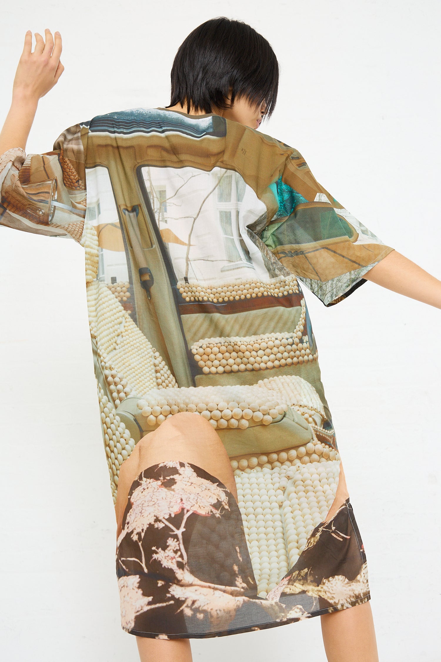 A person standing with arms raised slightly, wearing an oversized Bless No. 77 Sauna Rider Holiday Dress in Print featuring corn and landscape elements.