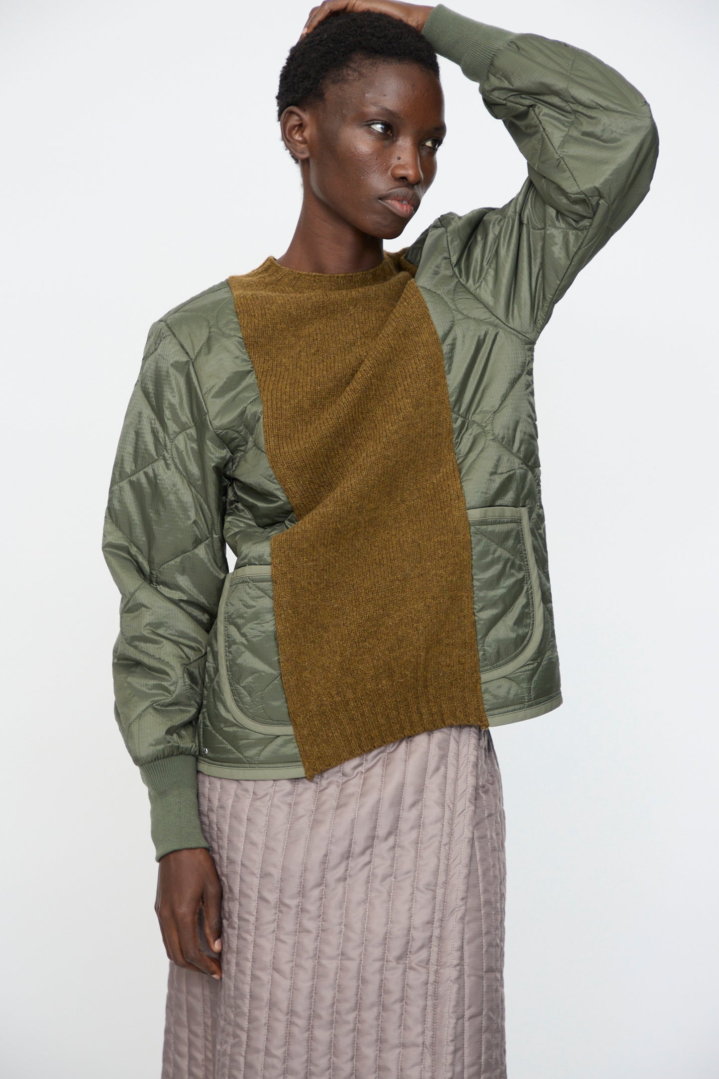 Model wears the Bless No. 78 Liningknitter Sweater in Olive, styled with a green quilted jacket featuring a brown panel and a light grey quilted skirt, standing against a plain white background.