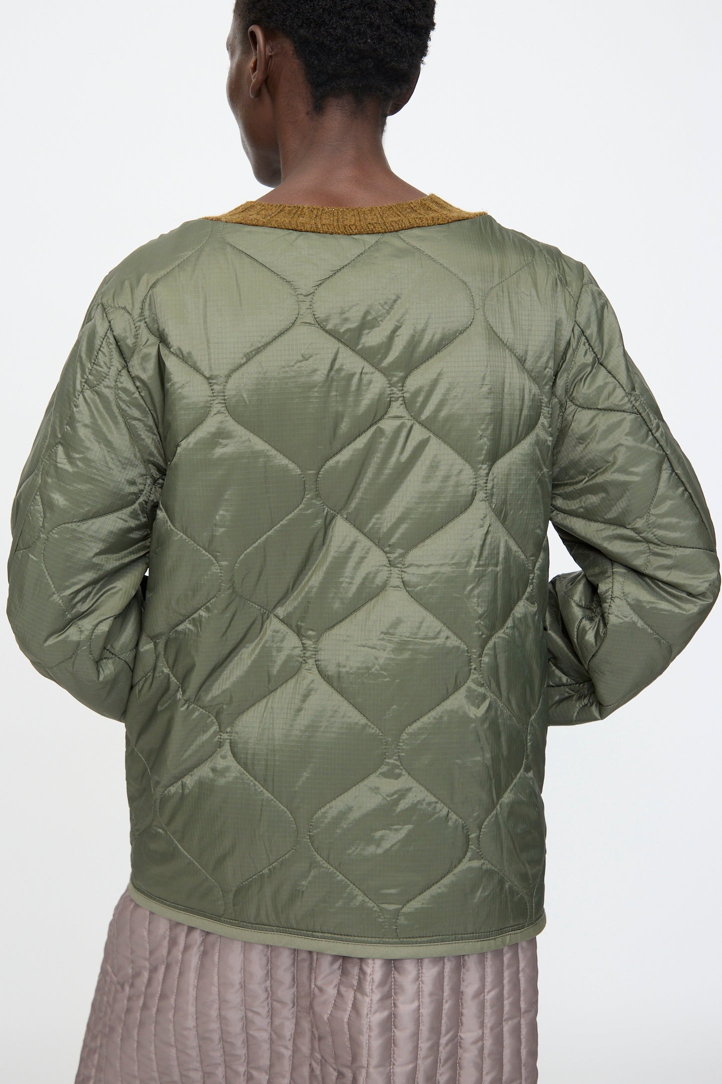 Person seen from the back wearing a quilted olive green jacket with a mustard collar, layered over the Bless No. 78 Liningknitter Sweater in Olive, paired with a light mauve quilted skirt.