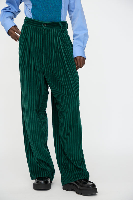 A person wears Bless’s No. 78 Ultrawidepleated Cord Pant in Dark Green, a light blue shirt, and black platform shoes, standing against a plain white background.