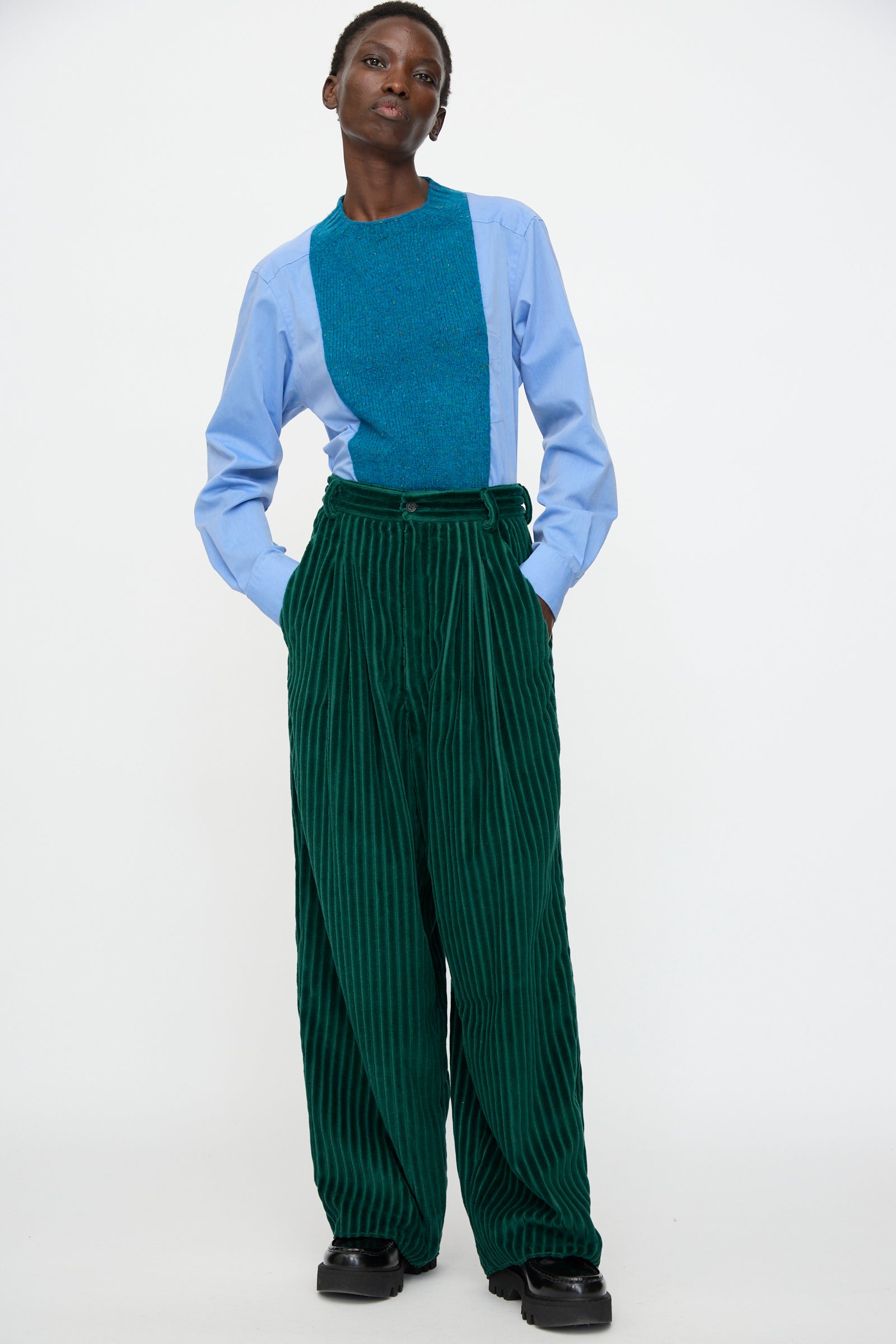 A person wears the Bless No. 78 Ultrawidepleated Cord Pant in Dark Green, paired with a light blue long-sleeve shirt featuring a teal textured panel, standing with hands in pockets against a plain white background.