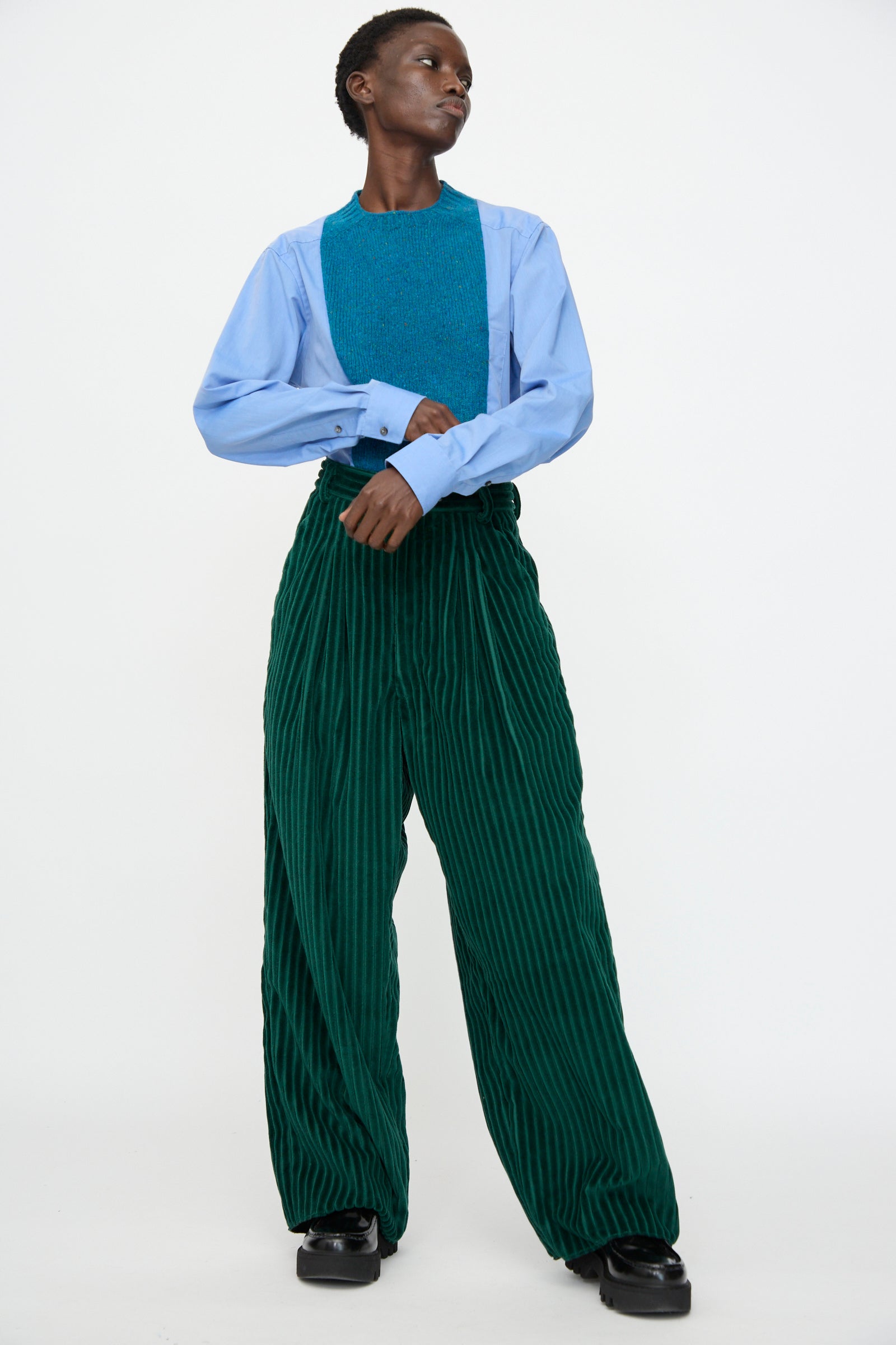 A person stands against a plain white background wearing a light blue shirt, teal textured vest, and Bless No. 78 Ultrawidepleated Cord Pant in Dark Green.