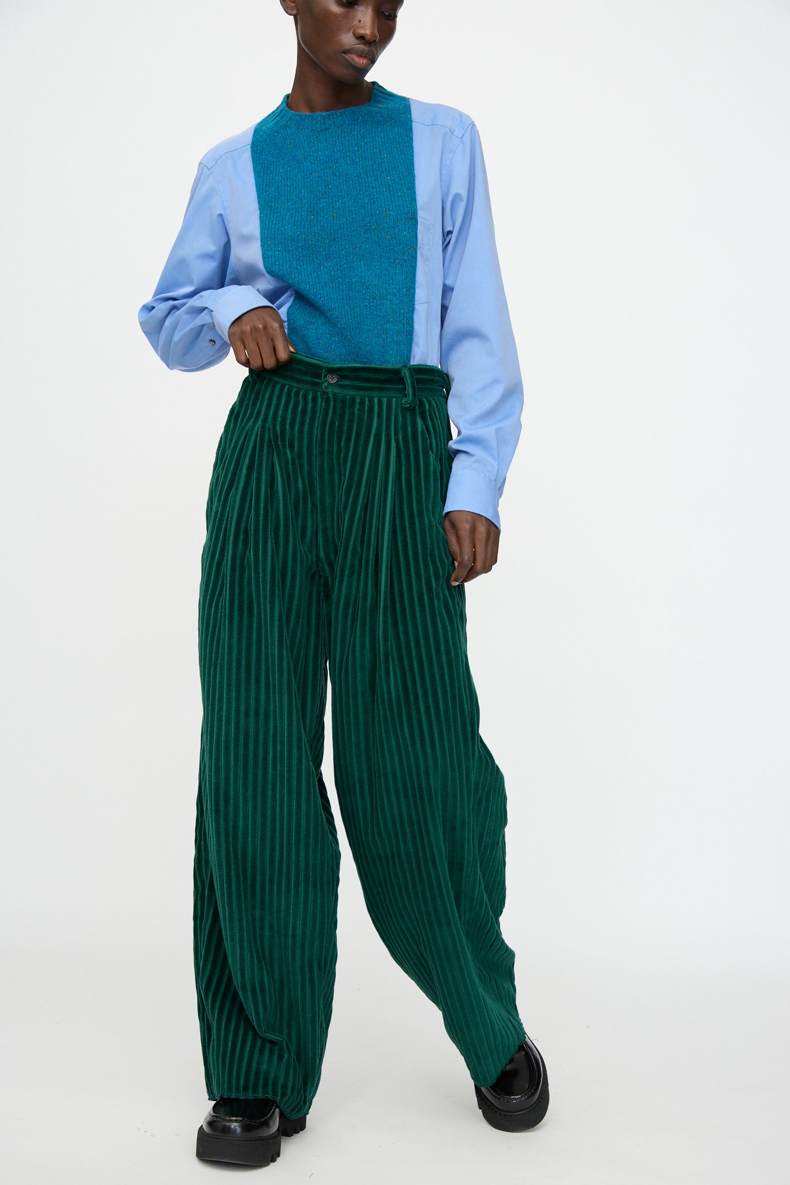 A person wears Bless's No. 78 Ultrawidepleated Cord Pant in Dark Green with a light blue shirt, teal textured vest, and black shoes, standing against a plain white background.