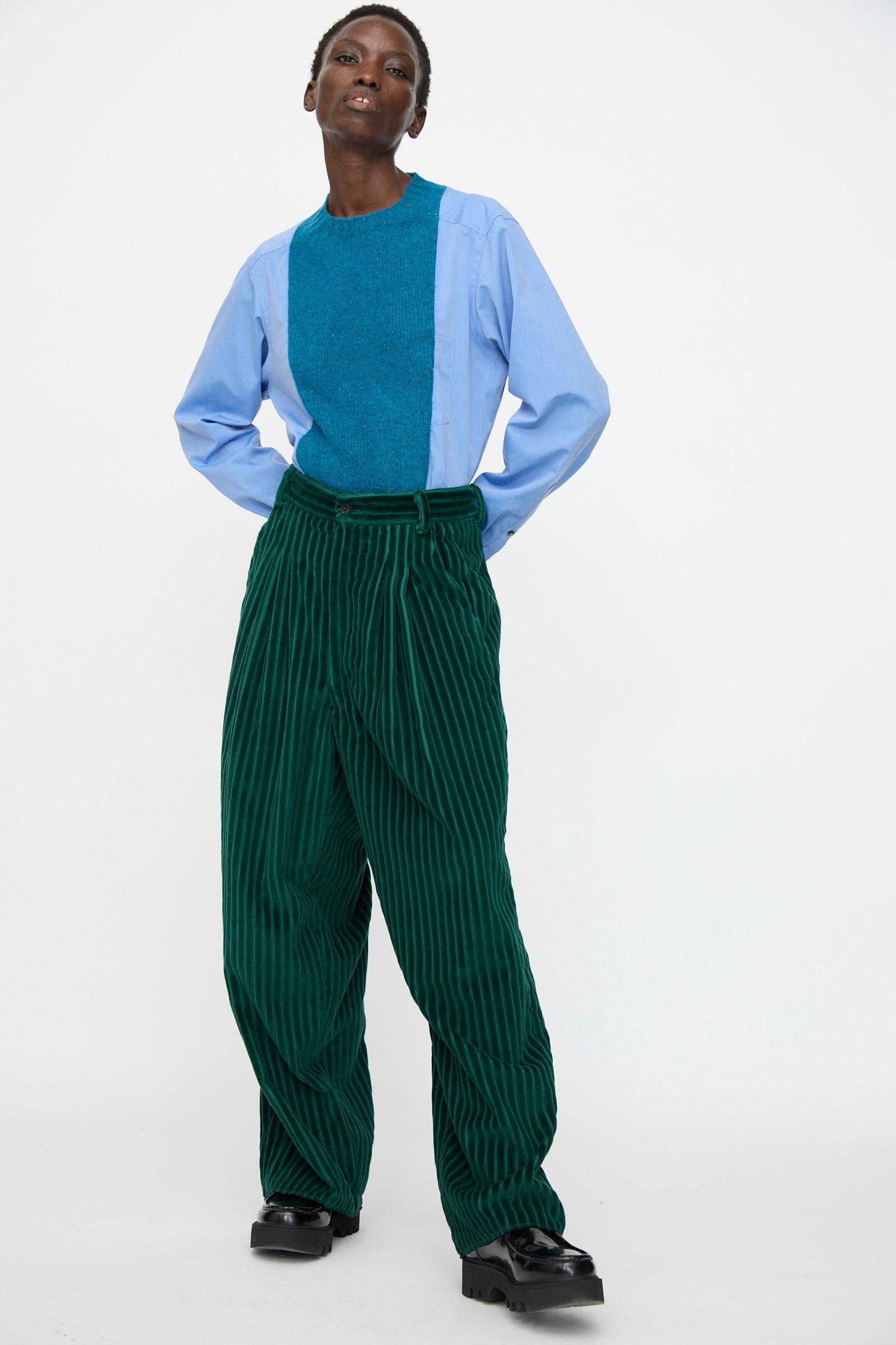 A person stands against a white background wearing a blue top, black shoes, and the Bless No. 78 Ultrawidepleated Cord Pant in Dark Green.