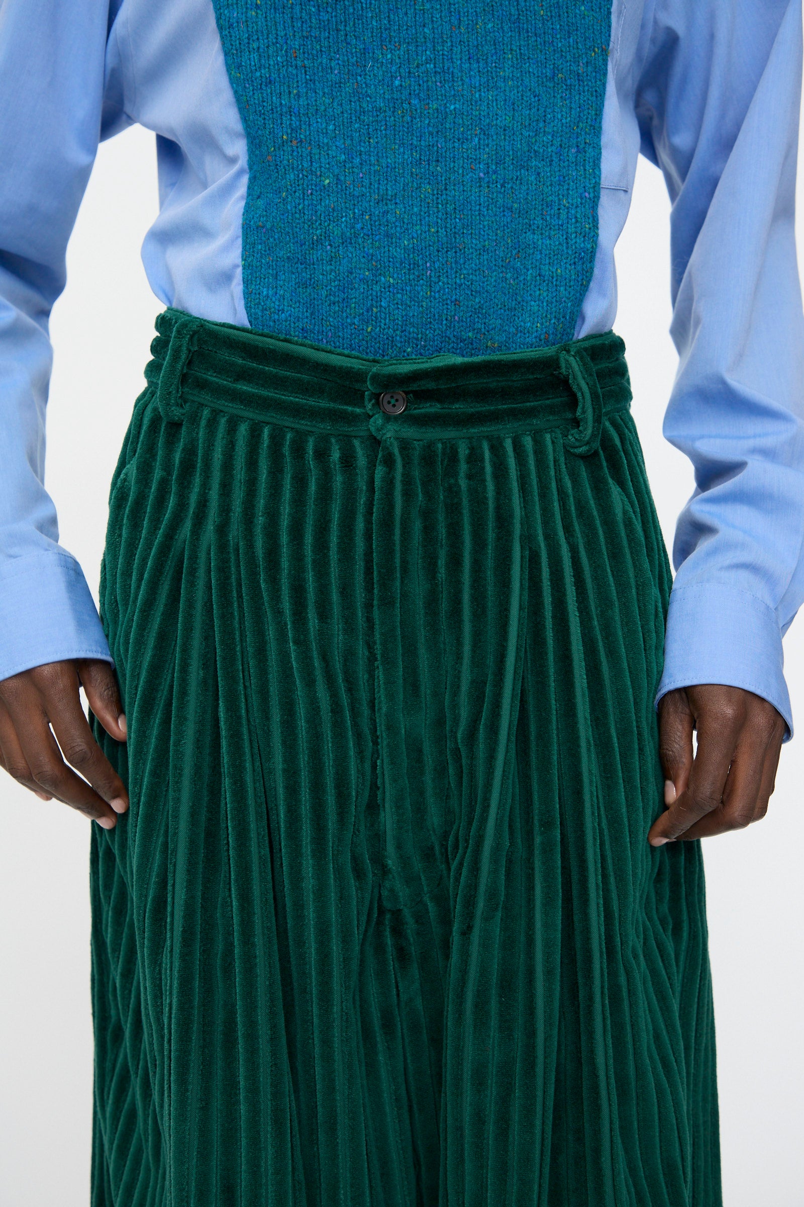A person wearing a light blue shirt, a blue-green textured vest, and the Bless No. 78 Ultrawidepleated Cord Pant in Dark Green.