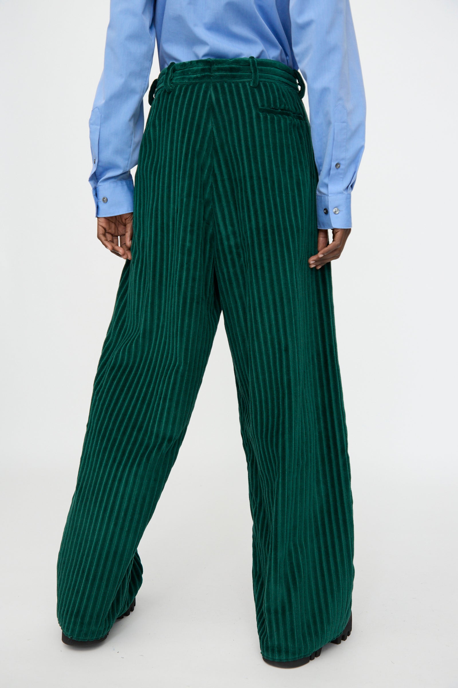 A person stands facing away from the camera against a plain background, wearing Bless’s No. 78 Ultrawidepleated Cord Pant in Dark Green and a light blue button-up shirt.