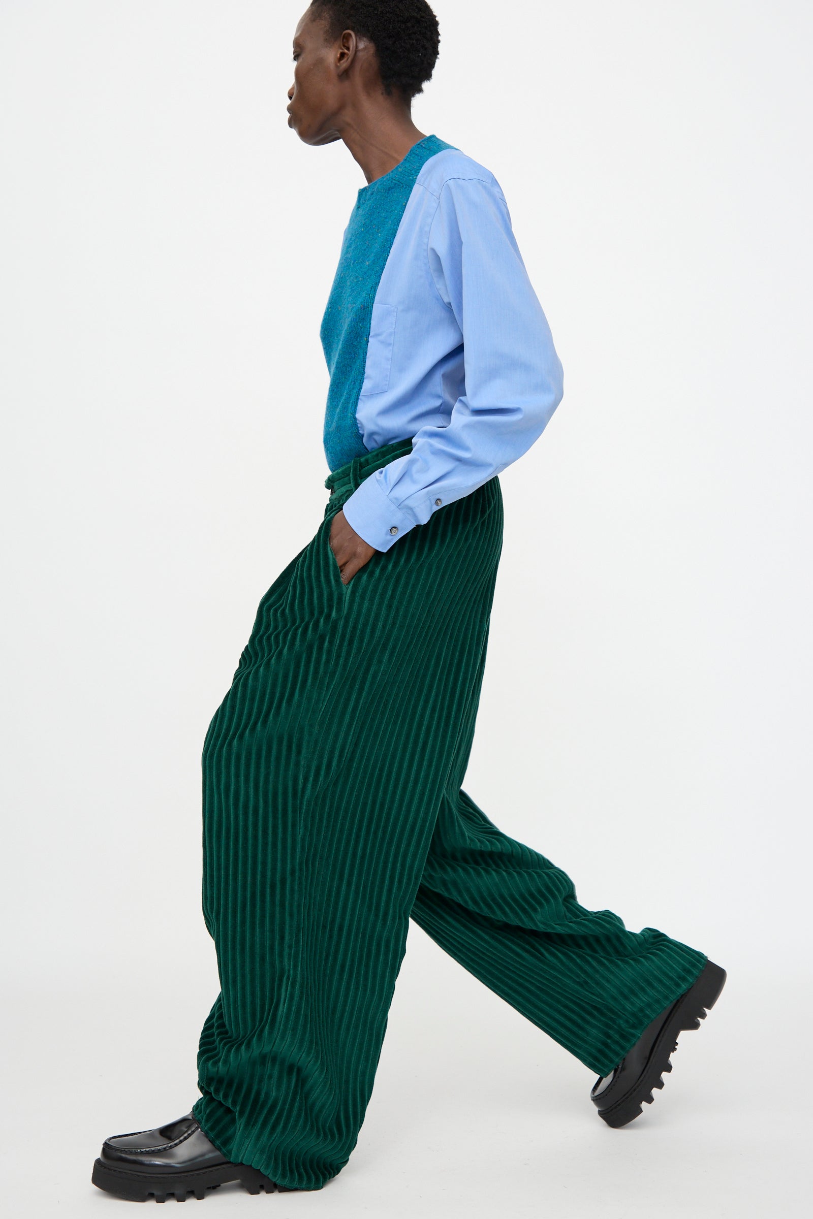A person walks in profile against a white background, wearing a light blue shirt, teal textured vest, black chunky shoes, and Bless No. 78 Ultrawidepleated Cord Pant in Dark Green.