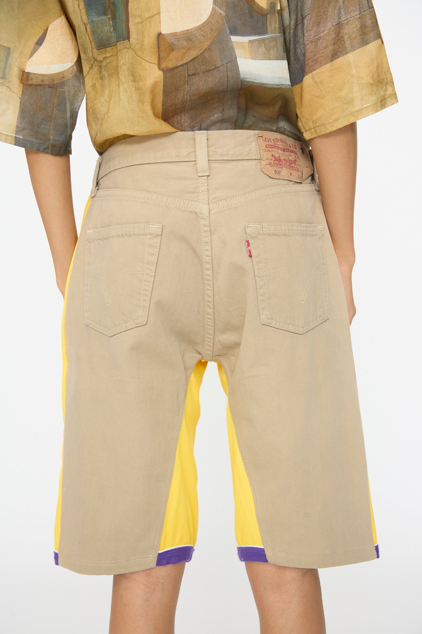Person shown from the back wearing Bless No. 81 Overjoggingshorts in Vintage Basketball Unique II—relaxed fit, knee-length beige shorts with yellow side panels—paired with a patterned shirt, set against a plain background.