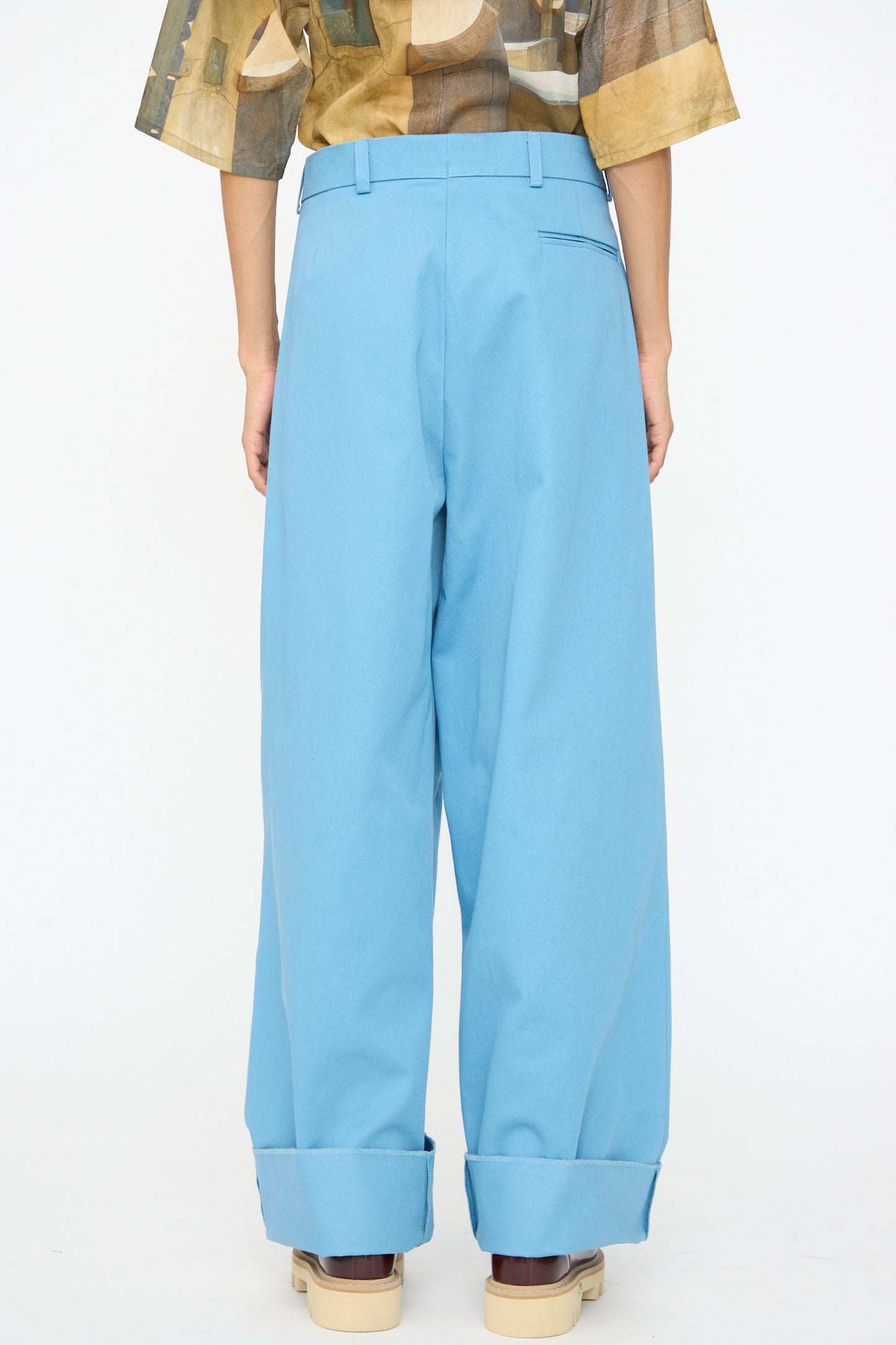 A person stands facing away, wearing the Bless No. 81 Ultrawidepleated Pant in Skyblue with cuffs and beige platform shoes, with part of a patterned shirt showing.