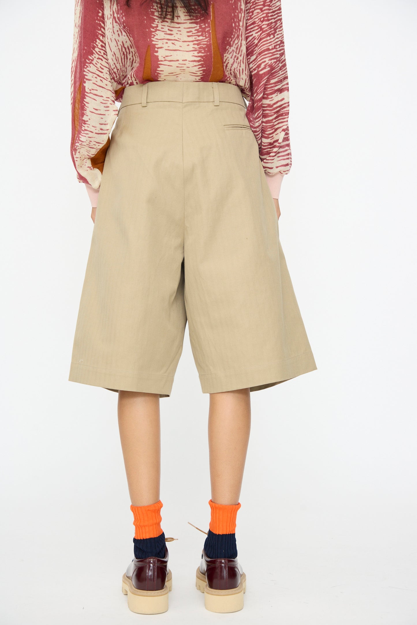 A person, seen from behind against a plain white background, wears a red patterned shirt, Bless No. 81 Ultrawidepleated Shorts in Sandkoeper, orange socks, and brown platform shoes.