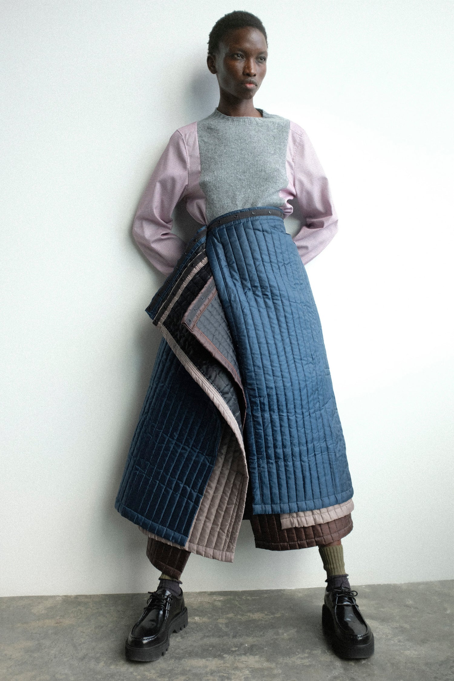 A woman wearing a long, quilted blue skirt with a light pink top against a white background.