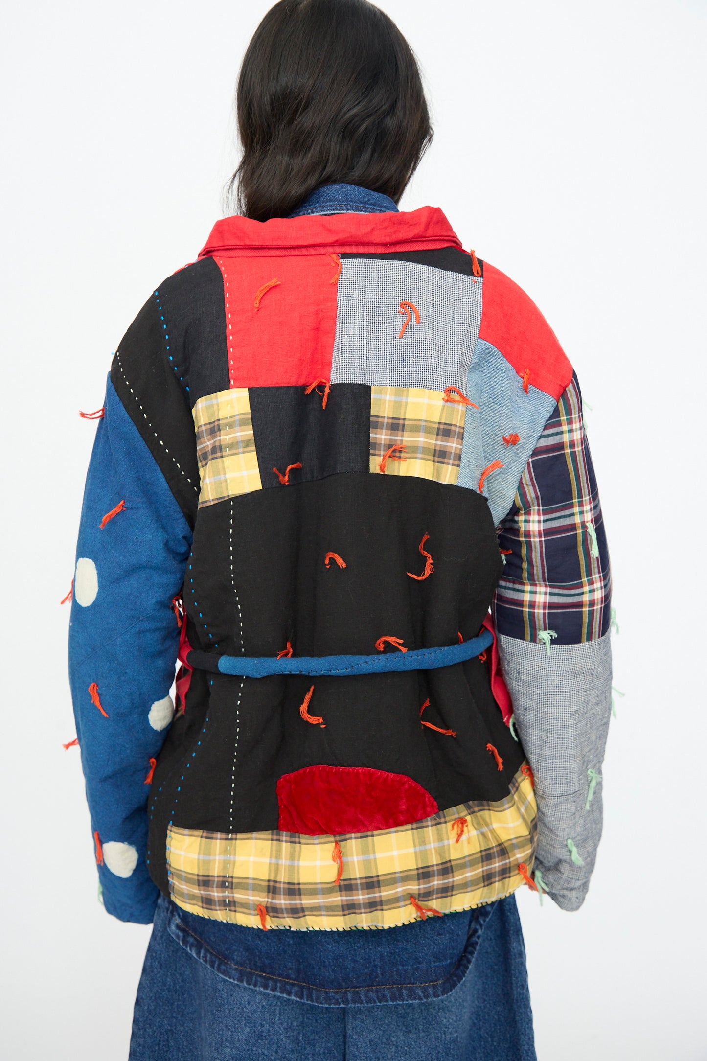 A person wears the CMC Polka Assembly Coat—a colorful, hand-quilted 100% cotton coat with mixed patterns and red stitching—paired with a denim skirt, photographed from behind. Thoughtfully made in New York.