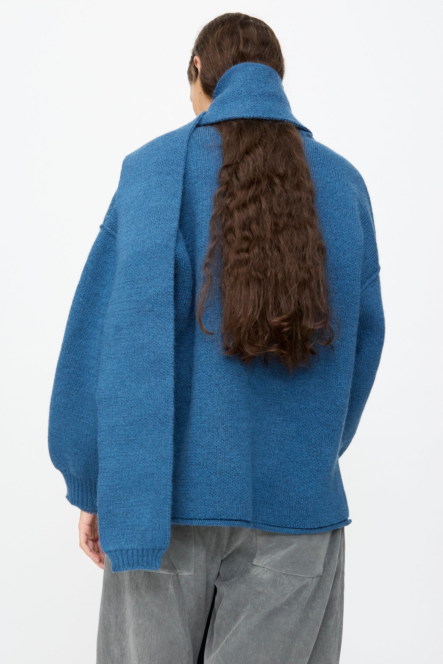 A person with long brown hair is seen from the back against a plain light background, wearing the Cawley British Merino Wool Long Scarf in Sea draped over a blue sweater and gray pants.