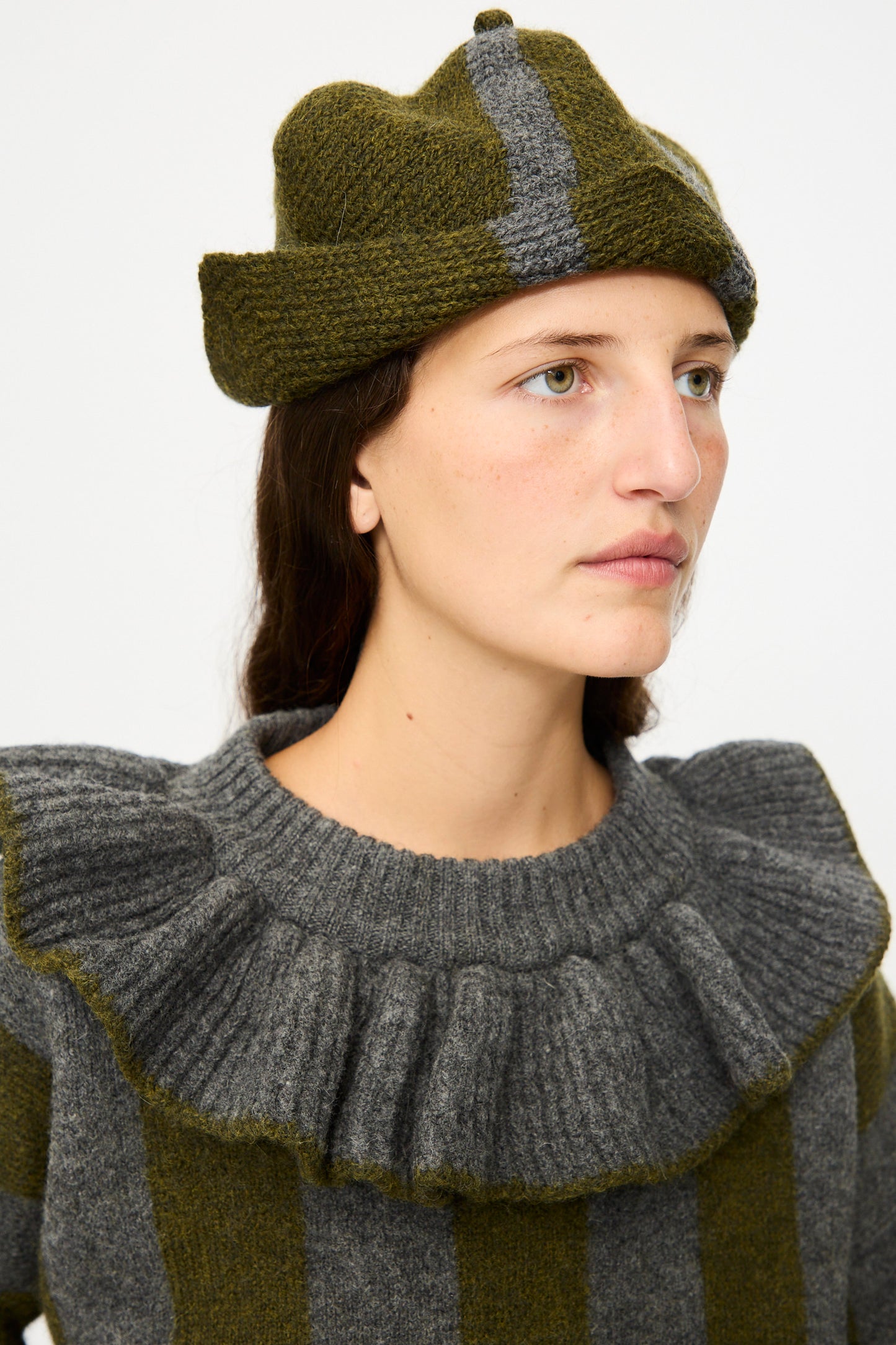 A person wearing a gray and olive green striped sweater with ruffled shoulders and the Cawley British Merino Wool Ski Hat in Moss and Slate, made in London, featuring vertical stripes, looks to the side.