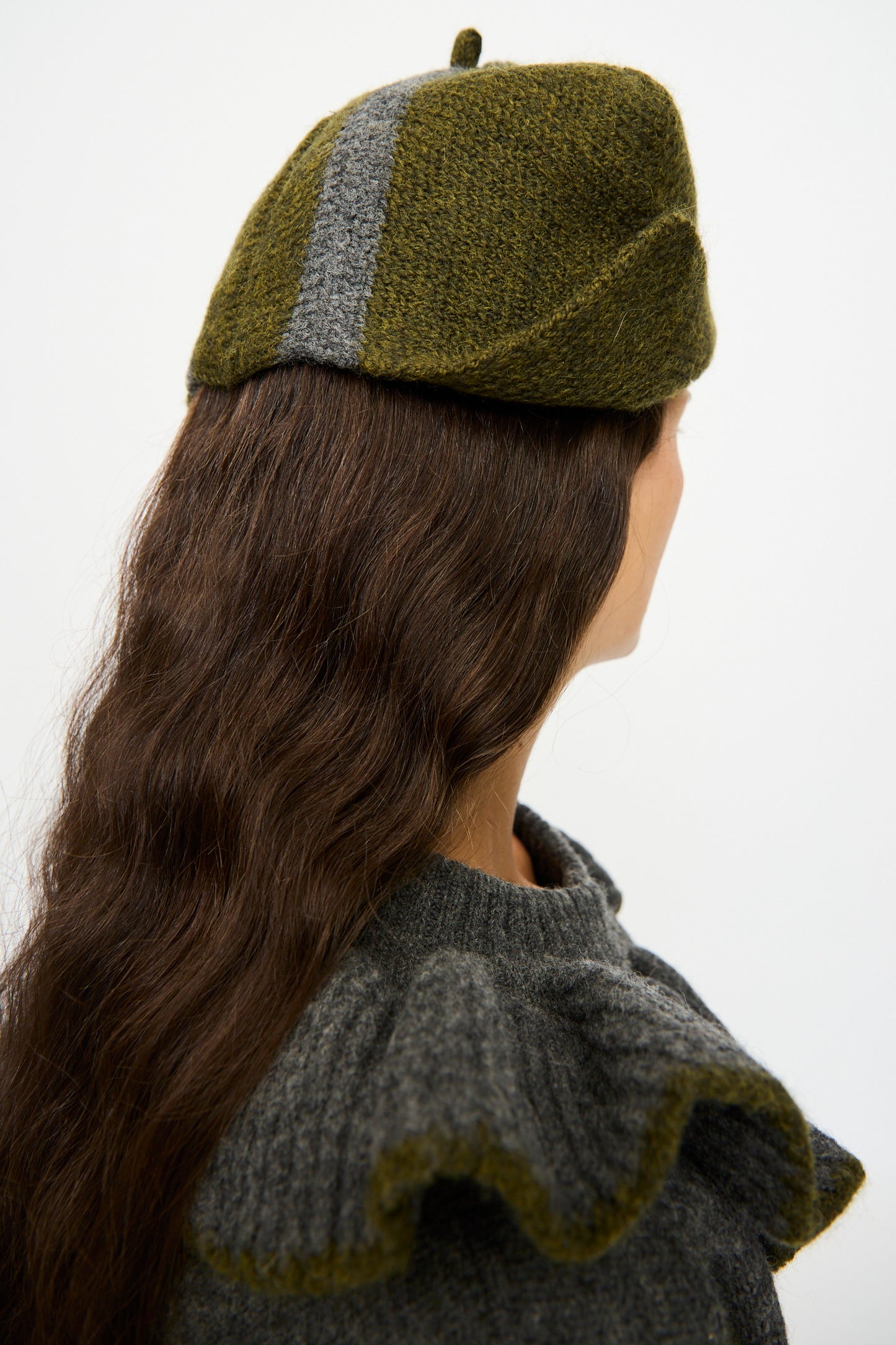 A person with long brown hair, facing away from the camera, wears Cawley's British Merino Wool Ski Hat in Moss and Slate and a gray ruffled sweater.