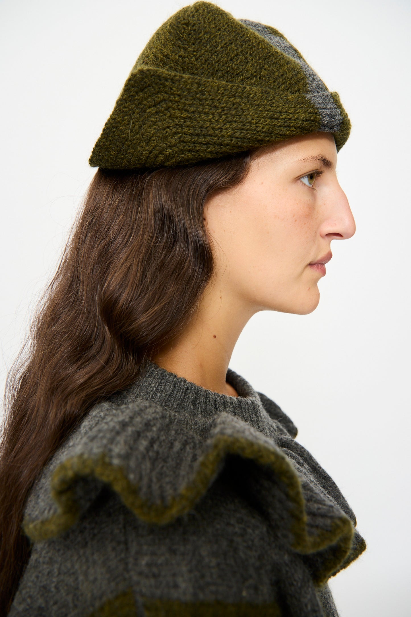 A person with long brown hair wears the Cawley British Merino Wool Ski Hat in Moss and Slate and a gray sweater with ruffled shoulders, shown in profile against a plain background.