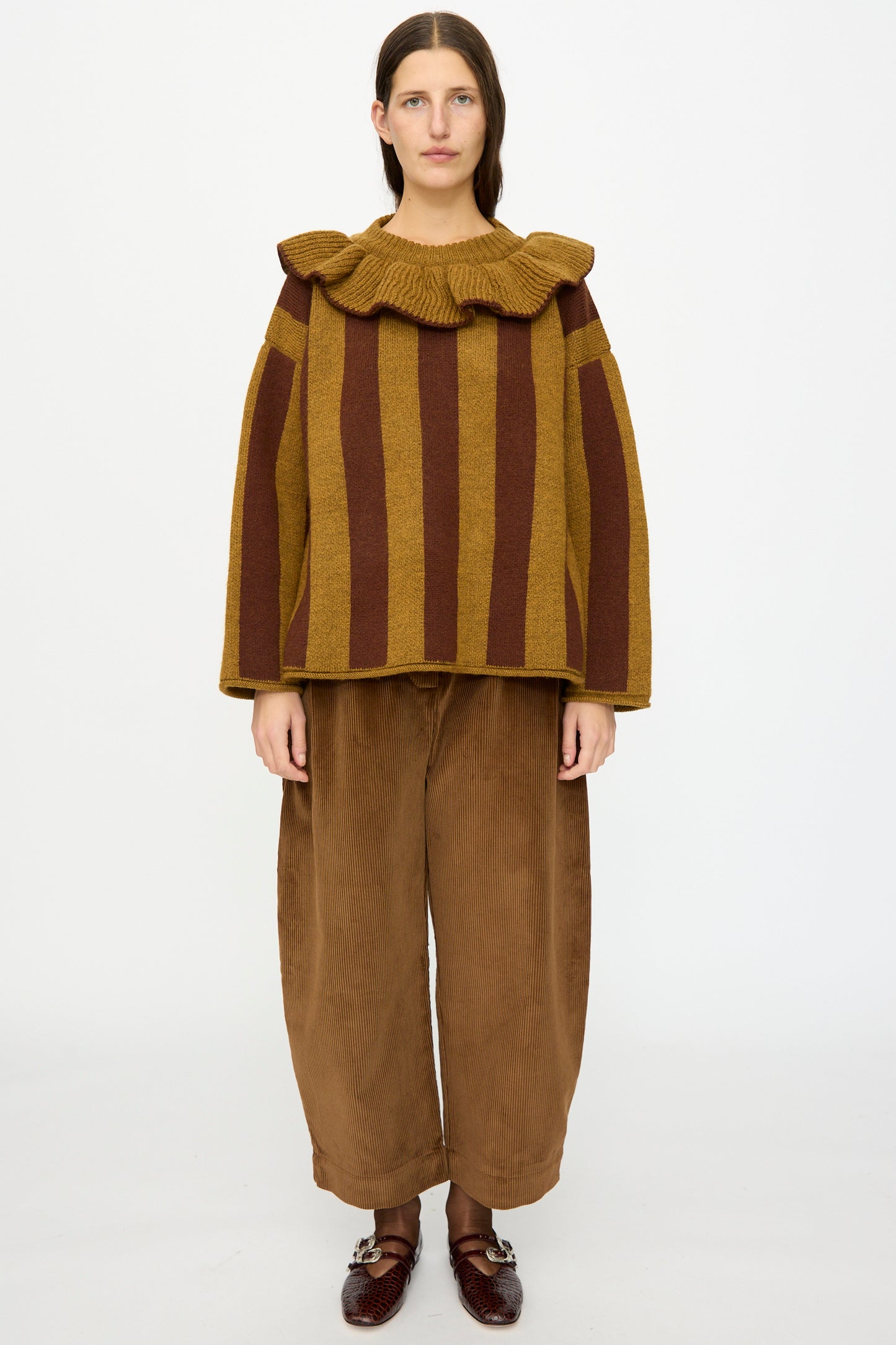 A person stands against a plain background, wearing the Cawley British Merino Wool Striped Ruffle Jumper in Goldfinch and Bordeaux, paired with wide brown pants and dark brown shoes.